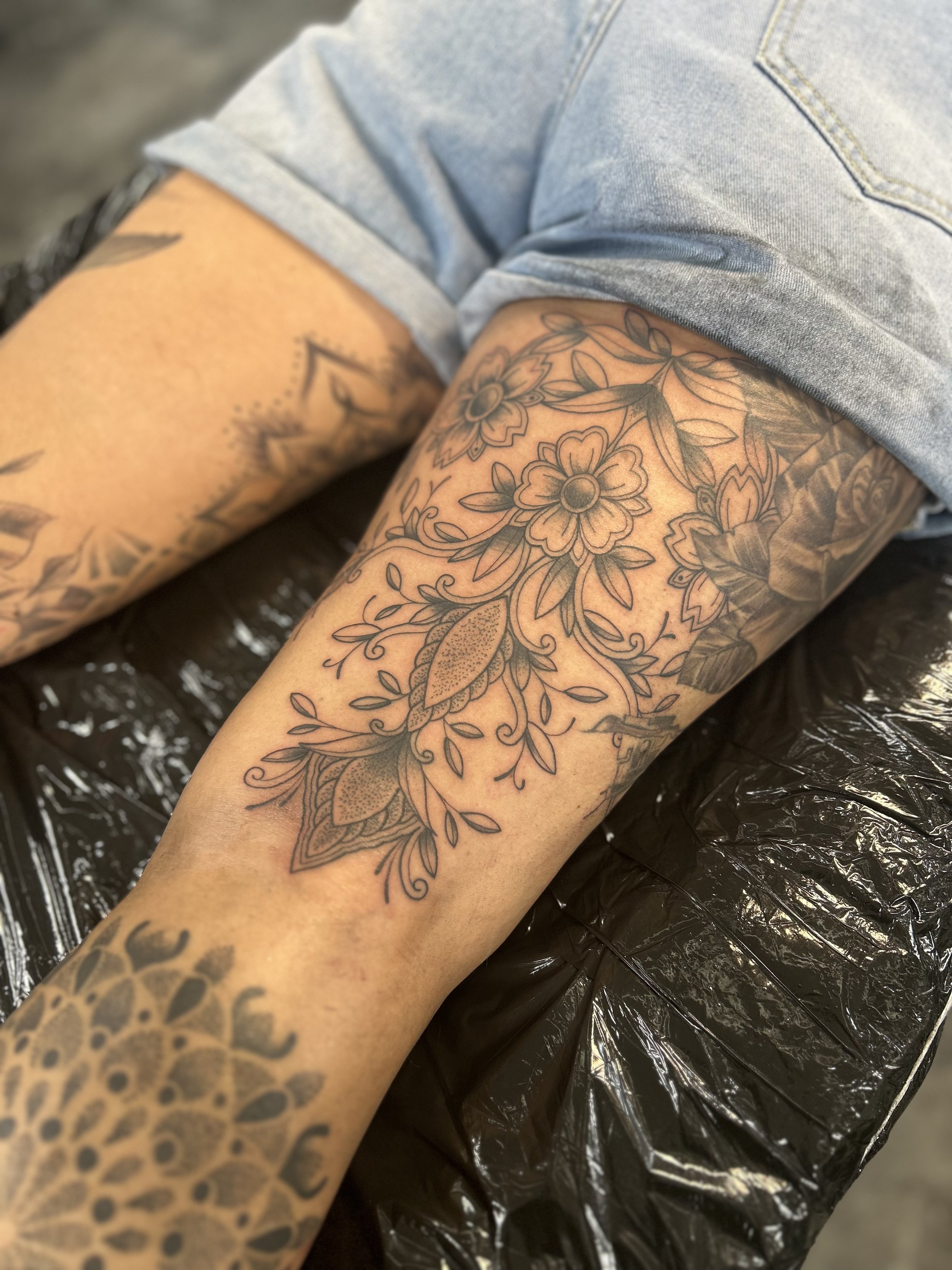 Close-up of a person's arm with intricate floral tattoo, wearing a light denim shirt, resting on a black plastic surface.