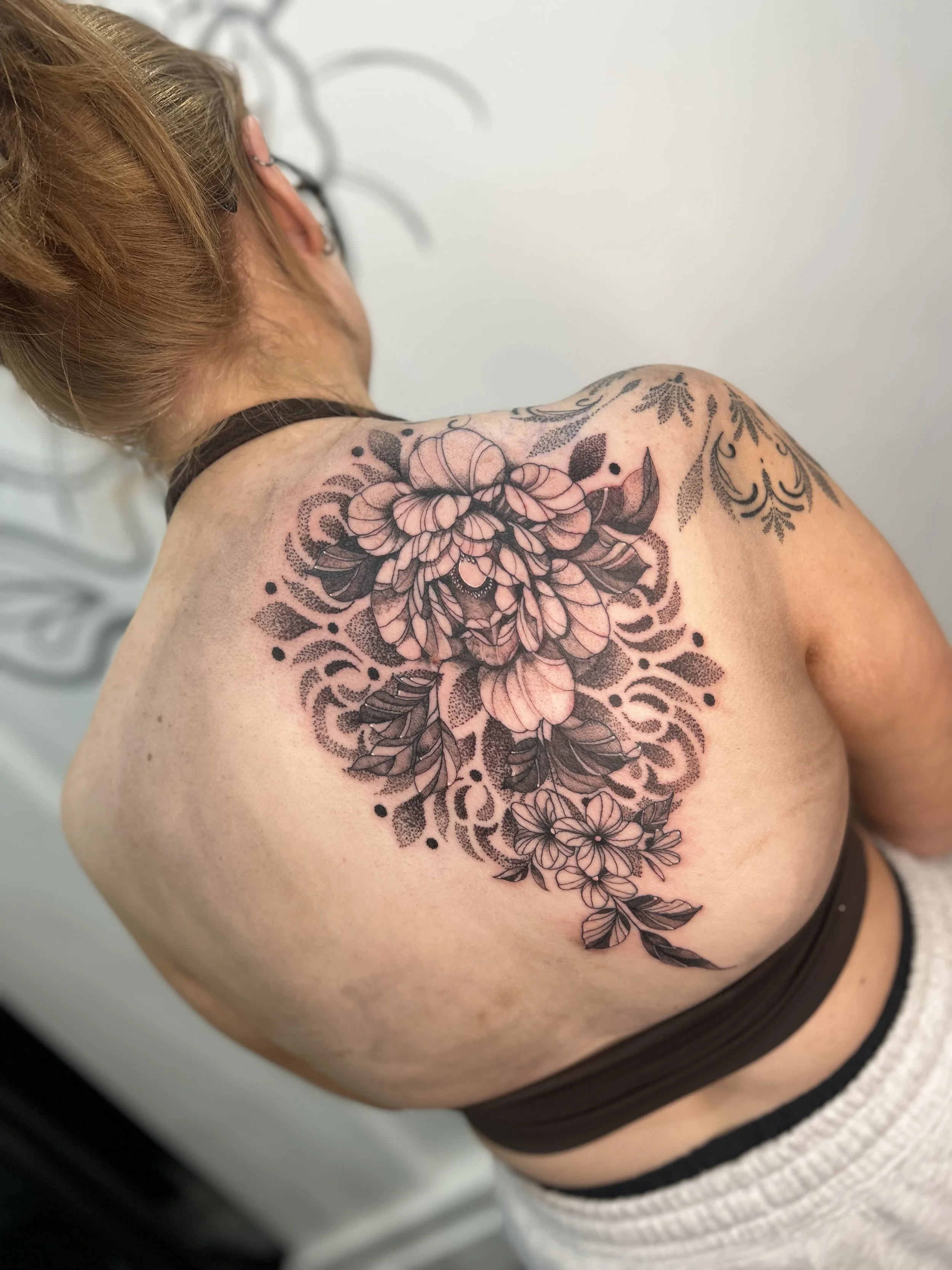 A person's shoulder and upper back with a large floral tattoo design in black ink, surrounded by decorative patterns. The person has blonde hair and is wearing a brown tank top and a white sweater.