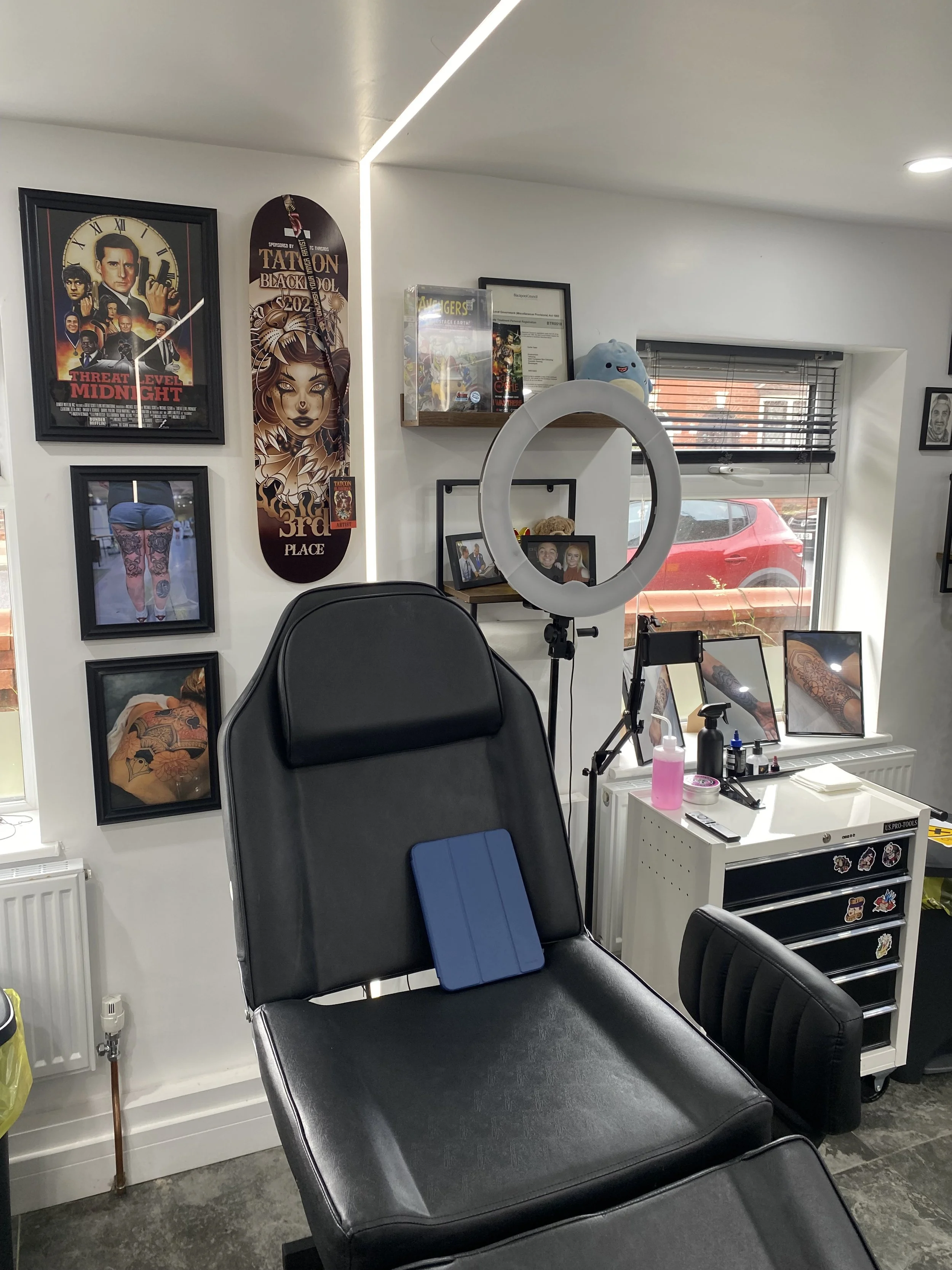 Tattoo studio with tattoo chair, framed artwork, a ring light, window, and office supplies.