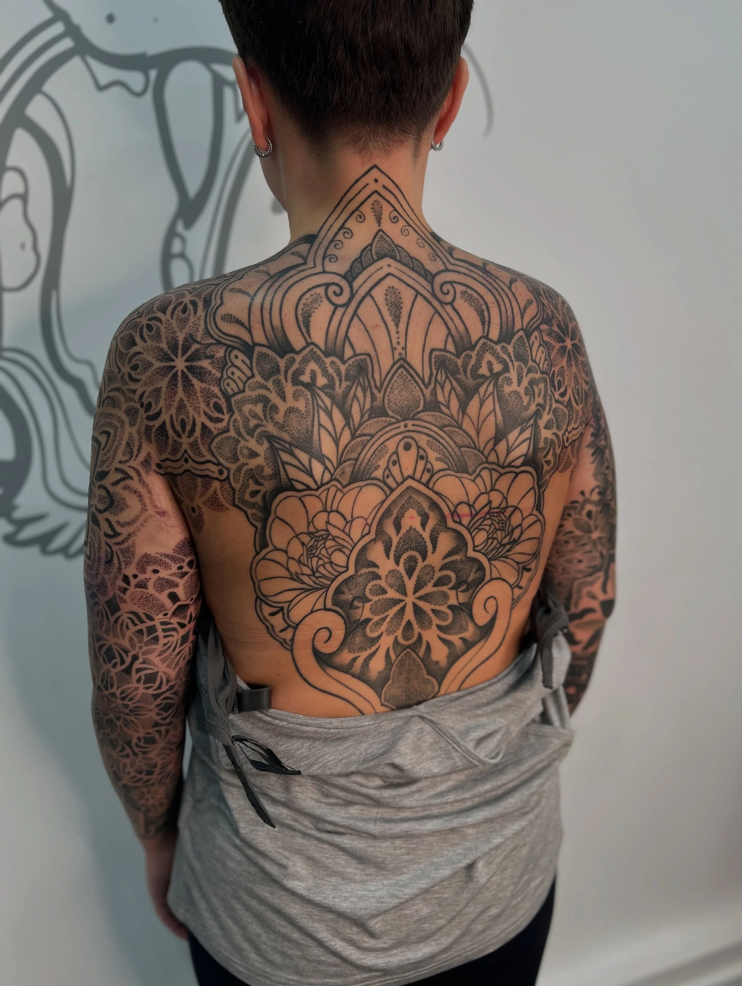 Back of a person's upper body with intricate black tattoos covering the shoulders, upper back, and arms, featuring floral and geometric patterns. The person is wearing a gray shirt and gray sweatpants with a black drawstring, standing against a plain