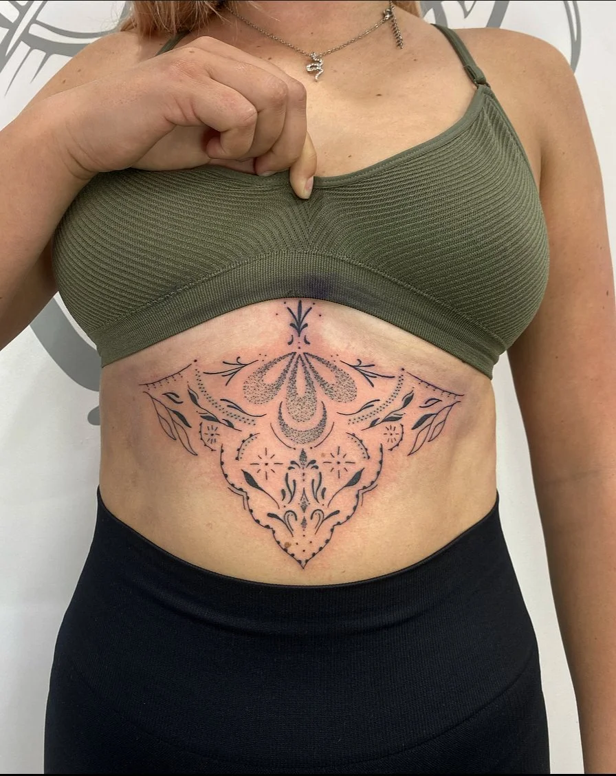 A woman in a green sports bra shows a fresh, intricate black tattoo on her abdomen featuring symmetrical decorative elements, dots, and floral patterns.