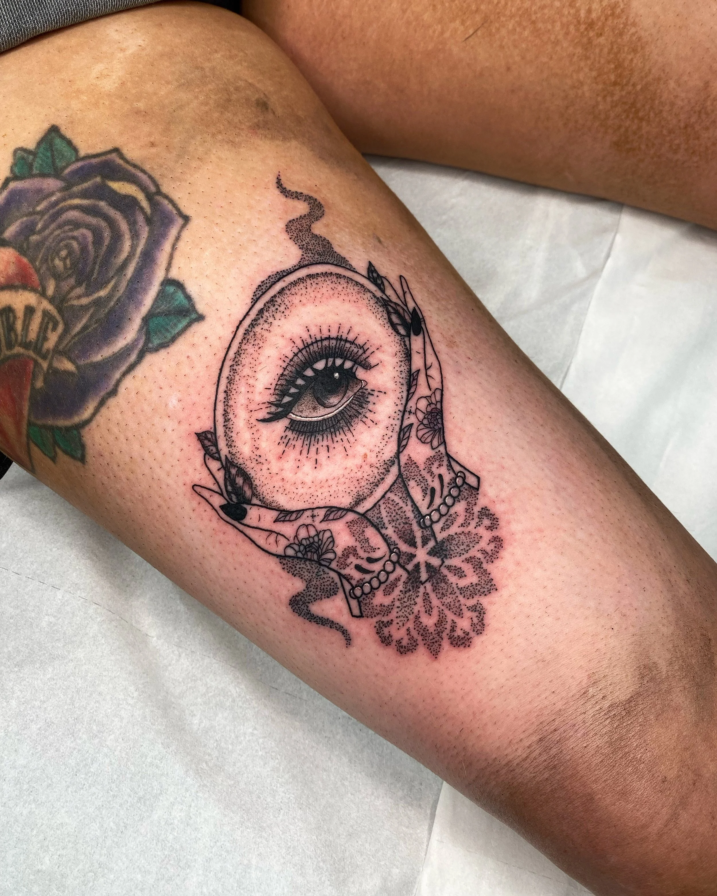 Tattoo of a detailed eye with lashes, surrounded by a circle with leaves and beads, and floral designs beneath it, on a person's forearm.