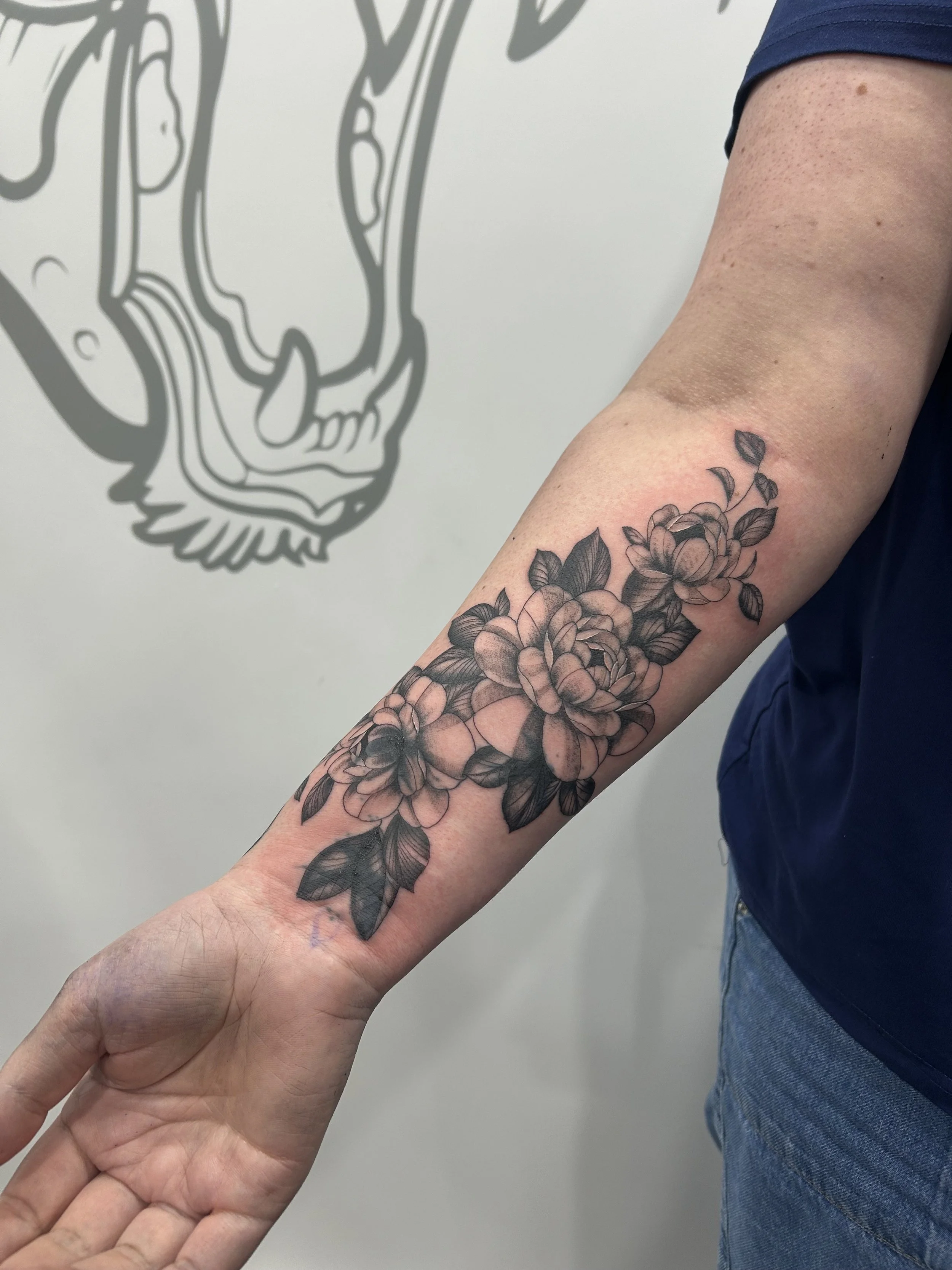A tattoo of flowers on a person's forearm, with a person standing nearby wearing a dark shirt and jeans, in front of a wall with a large outlined animal face design.