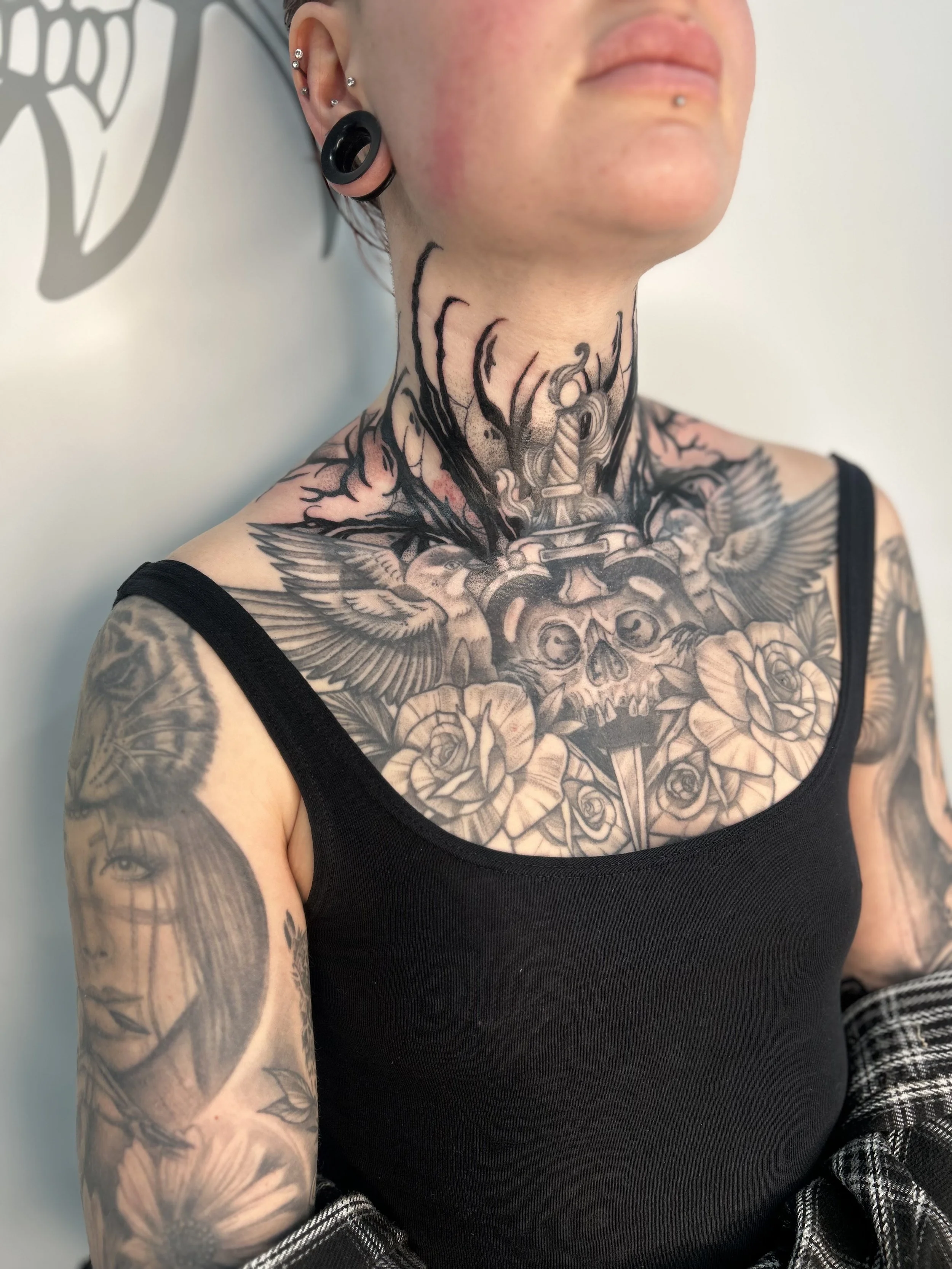 Close-up of a woman with tattoos on her neck and arms, wearing a black tank top and multiple earrings, including stretch gauge and several piercings on her ear, with a shadow profile of her face and part of her shoulder visible.