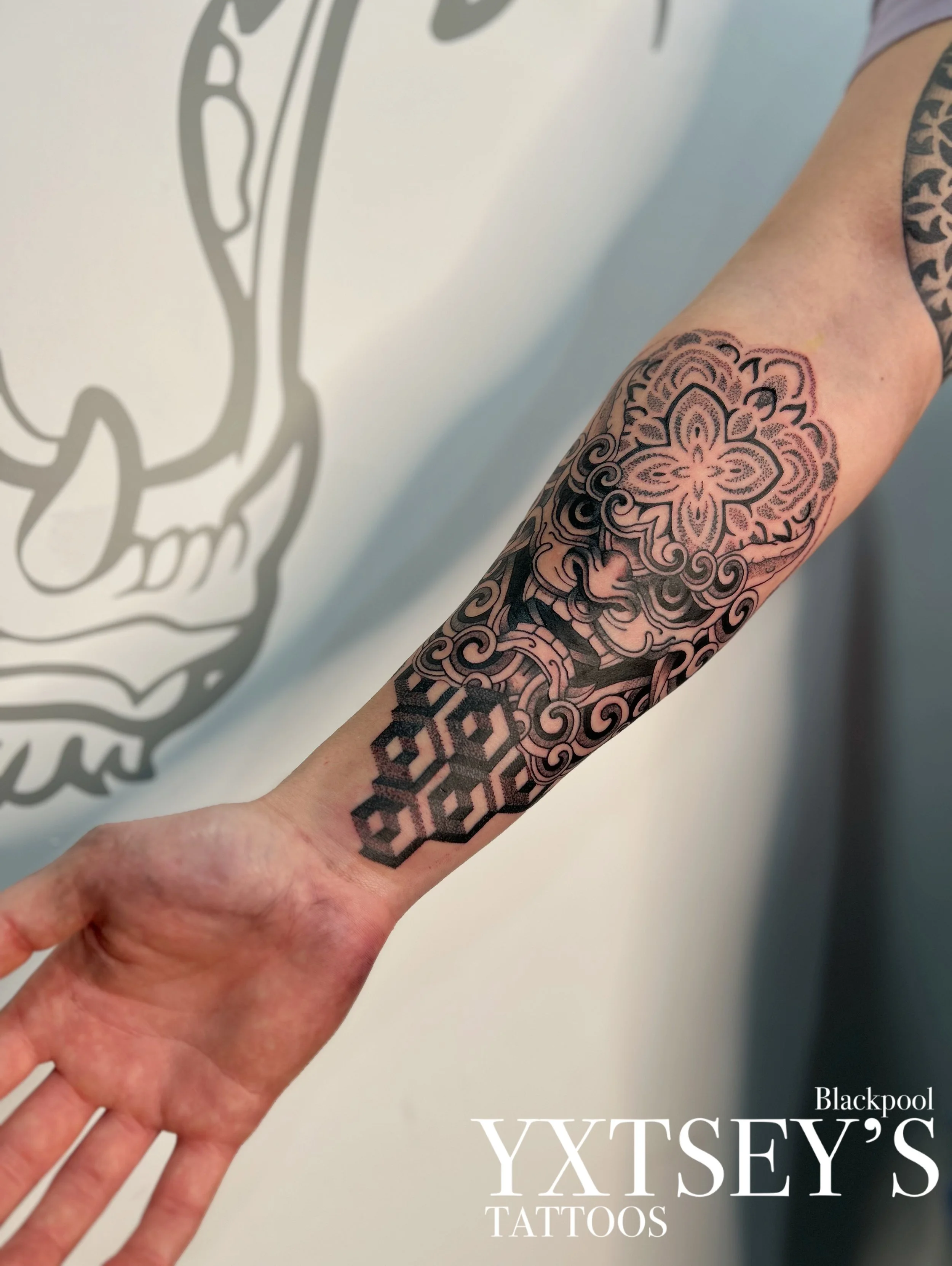 Close-up of an arm with a black and gray tattoo featuring floral and geometric patterns, with a blurred sign in the background that reads "Yxtsey's Tattoos" and part of a larger design.