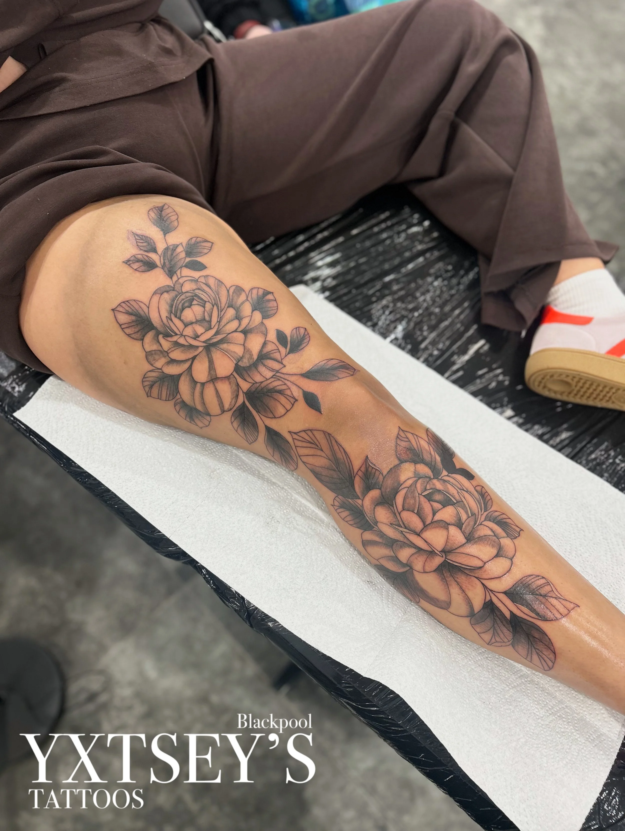 A person's forearm with a large, detailed black and gray floral tattoo, showing roses and leaves. The person is sitting in a tattoo parlor with a black bench and white paper covering the surface.