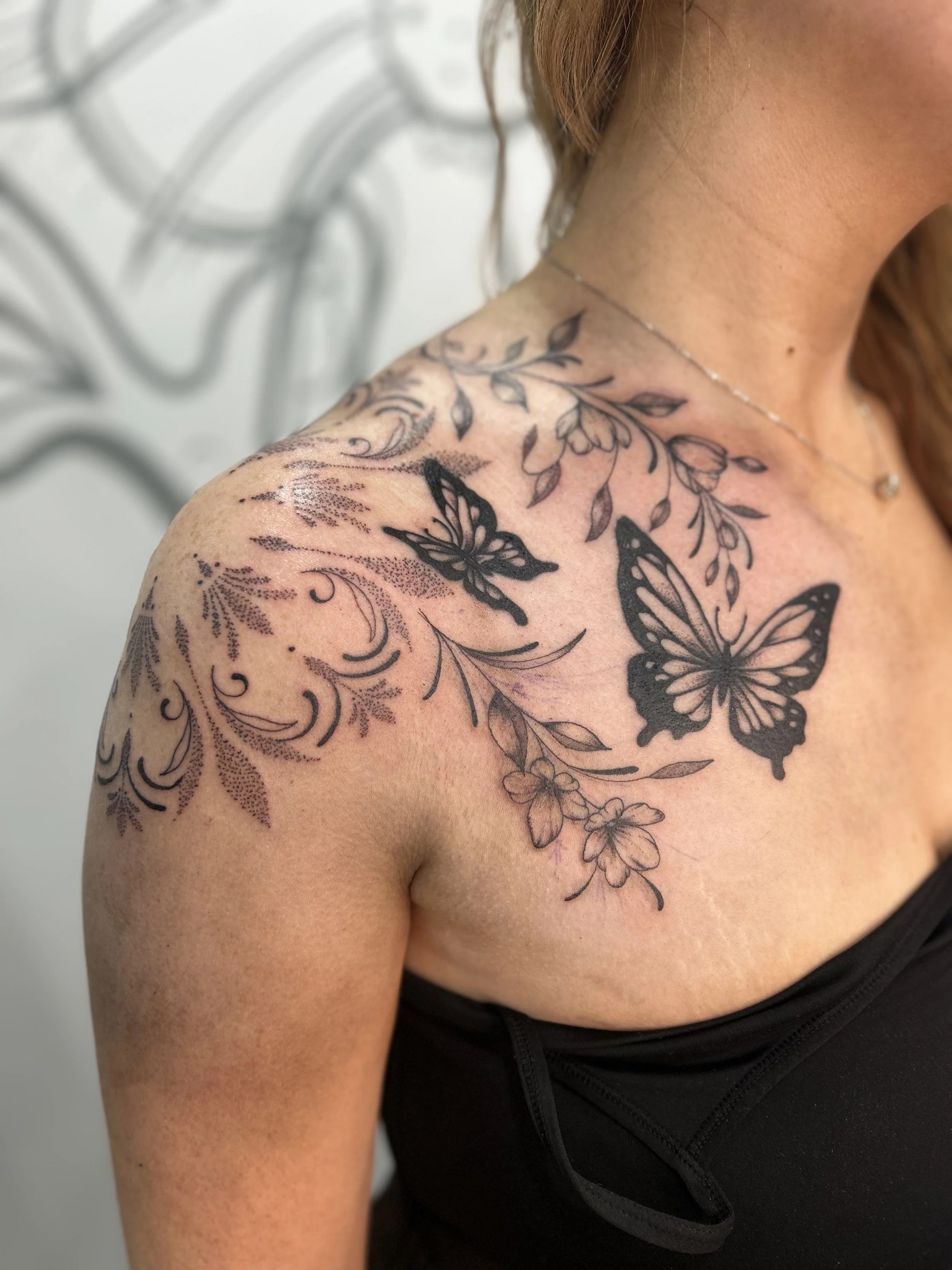 Close-up of a woman's shoulder and upper arm with a floral and butterfly tattoo, wearing a black athletic top and a delicate necklace, against a light background.