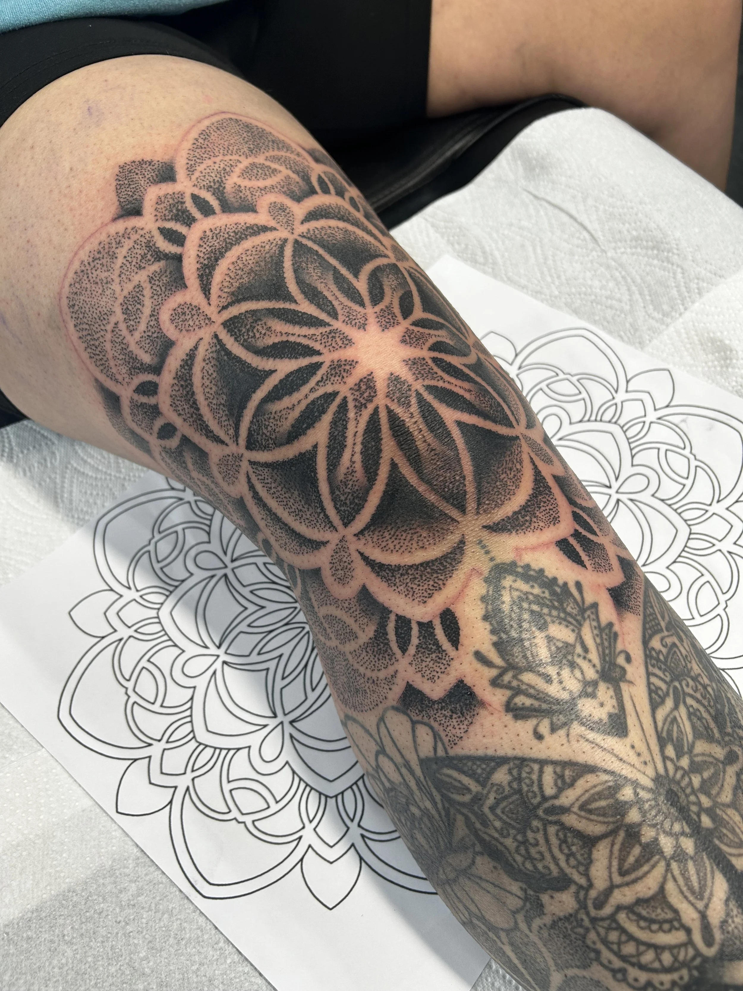 A person's arm with a large black and gray floral tattoo, resting on a tattoo design outline on white paper.