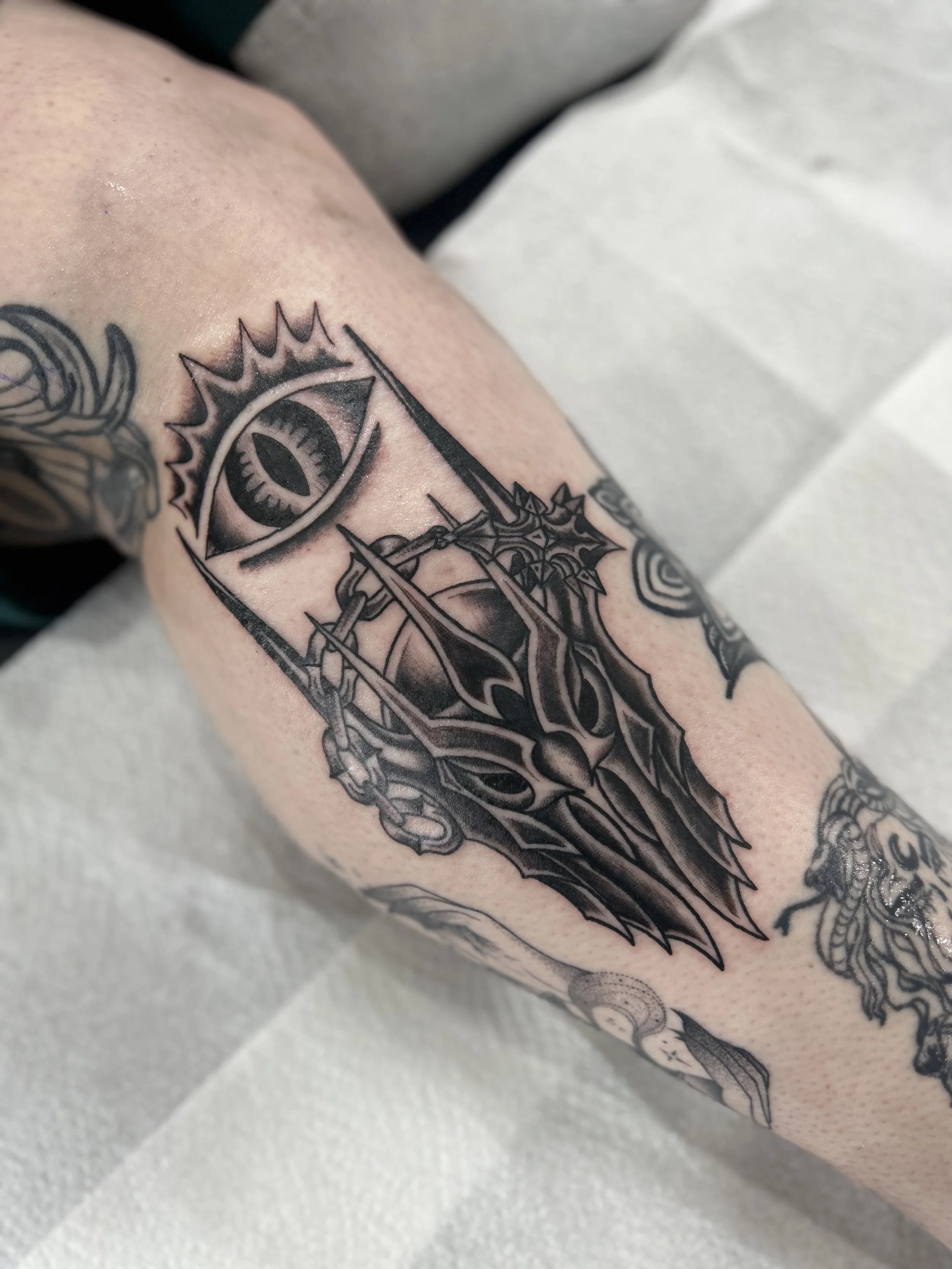 A close-up tattoo on a person's arm featuring a stylized eye within a geometric design, with chains and sharp, pointed elements surrounding it.