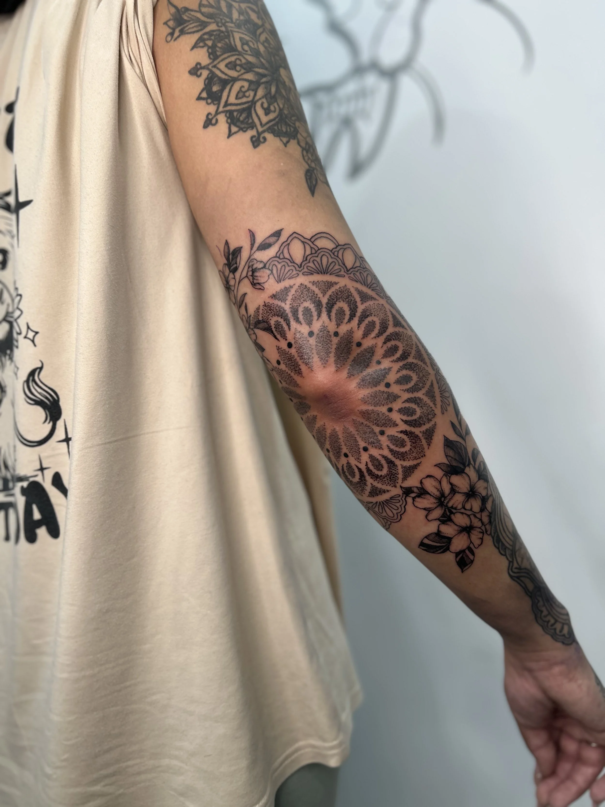 A person with detailed black and gray tattoos on their arm, including floral and geometric patterns, holding hands with another person. The person is wearing a beige sleeveless top.