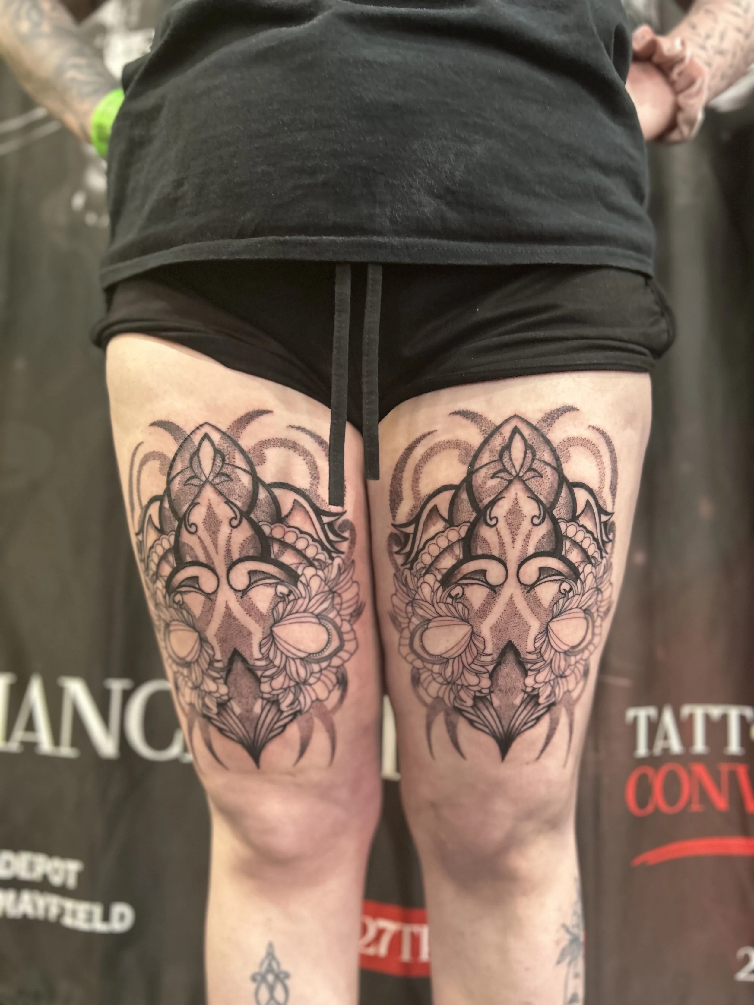 Close-up of a person's upper thighs tattooed with intricate black and gray floral designs, wearing black shorts and a gray T-shirt.
