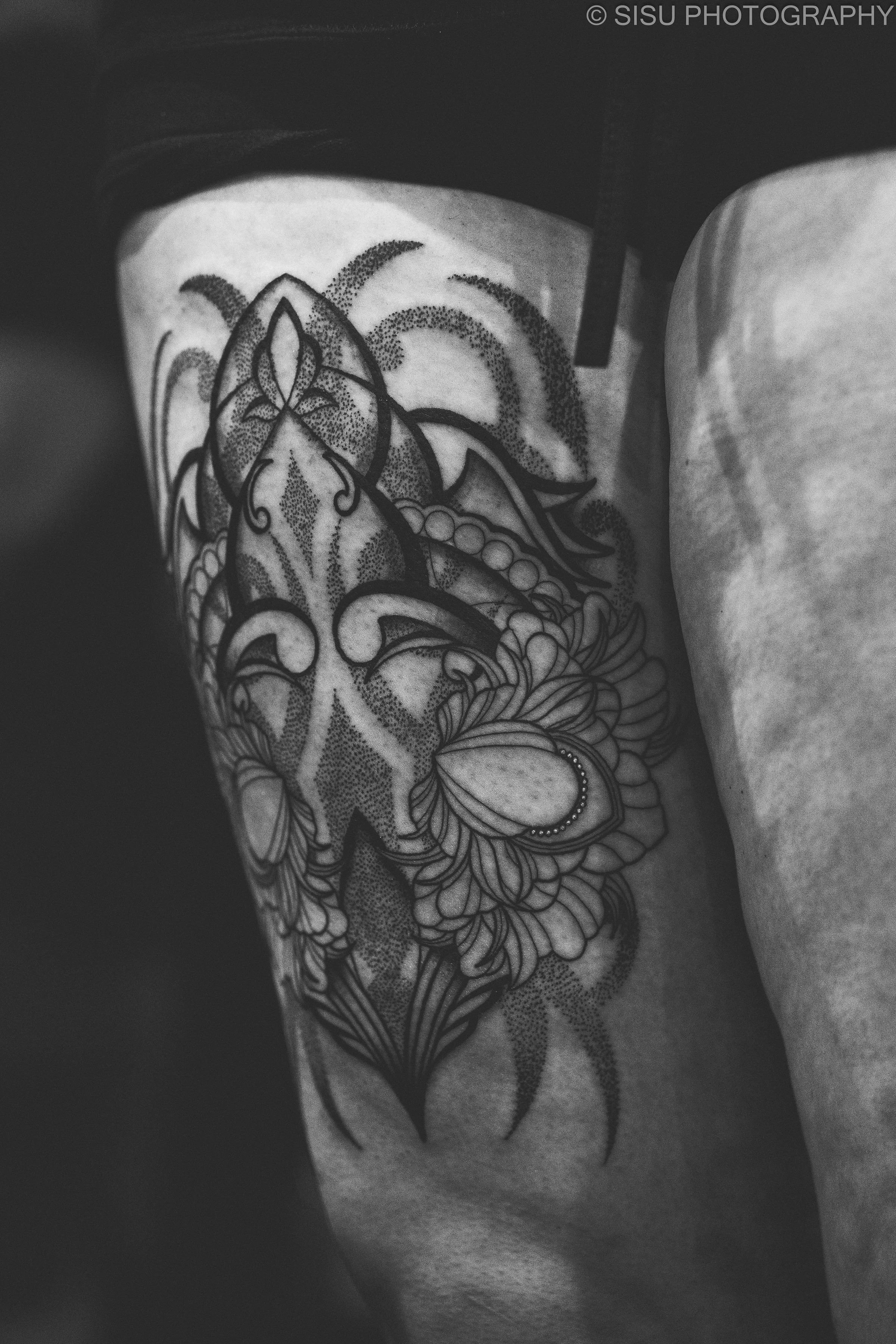 Black and white photo of a person's tattooed thigh featuring intricate floral and geometric patterns.