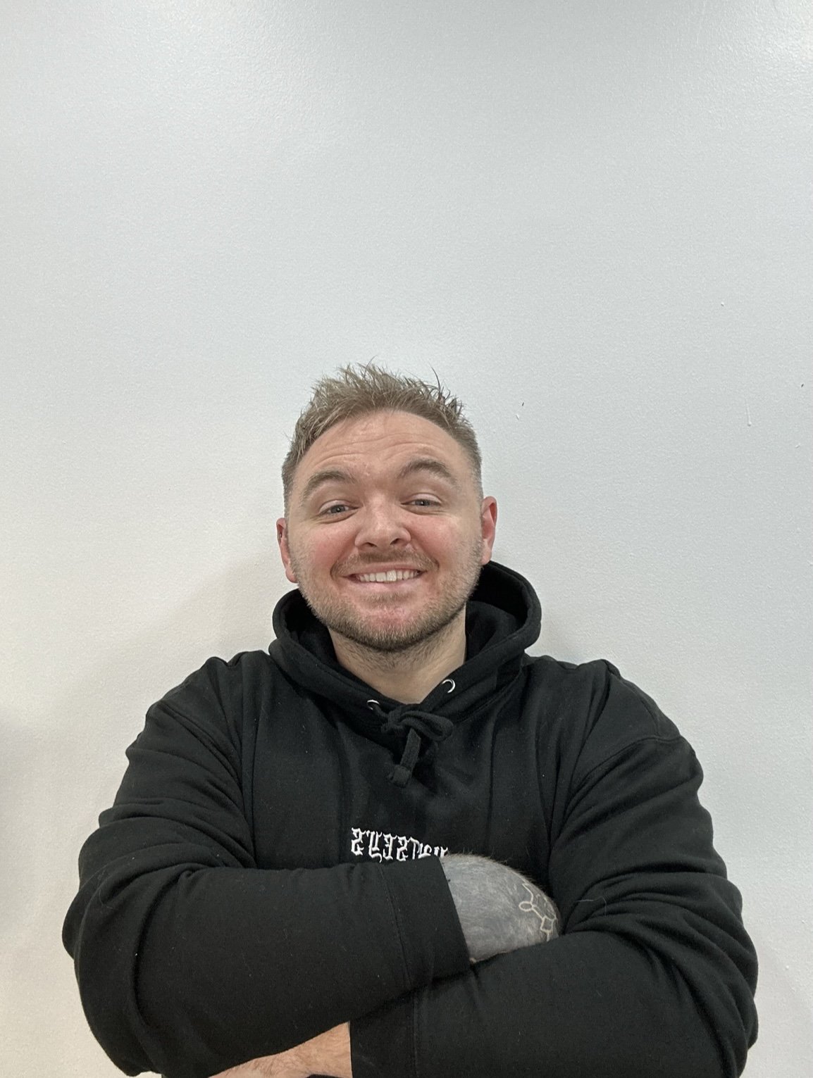 A smiling man with short light brown hair and a beard, wearing a black hoodie, standing against a plain white wall, holding a soccer ball.
