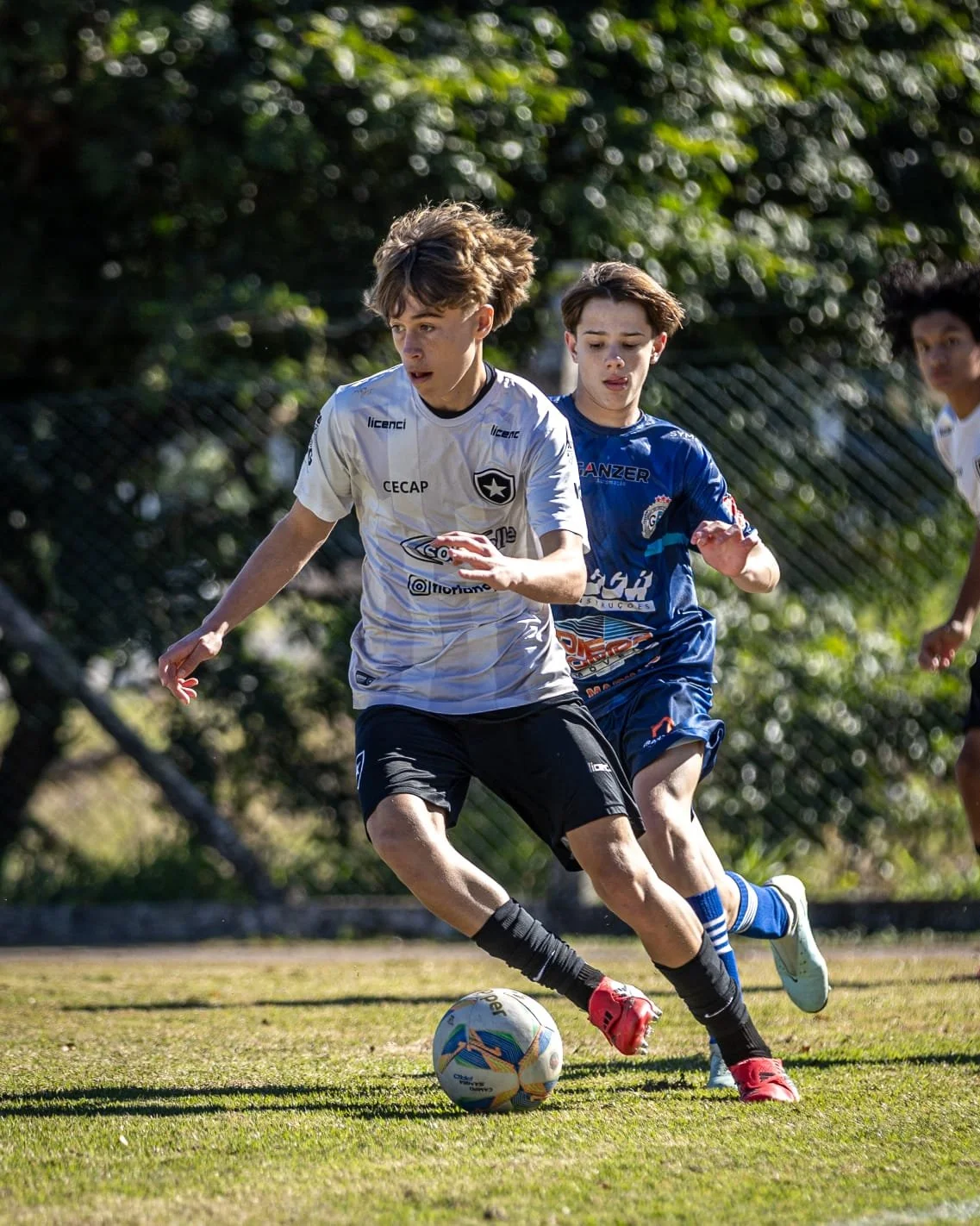 Two young boys playing soccer outdoors on a grassy field, with trees and a chain-link fence in the background. One boy in a white jersey with black shorts controls the ball, while the other in a blue jersey runs nearby.