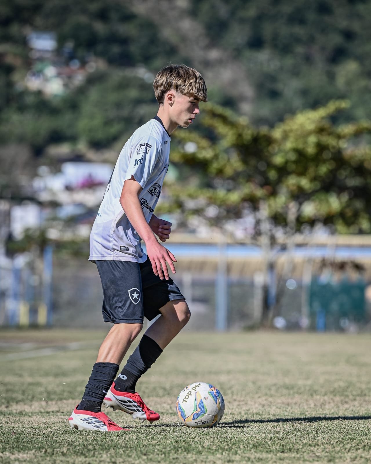A young male soccer player with light brown hair, wearing a grey jersey with logos, black shorts with a star emblem, red and black cleats, black knee-high socks, is about to kick a soccer ball on a grassy field with trees and blurred buildings in the