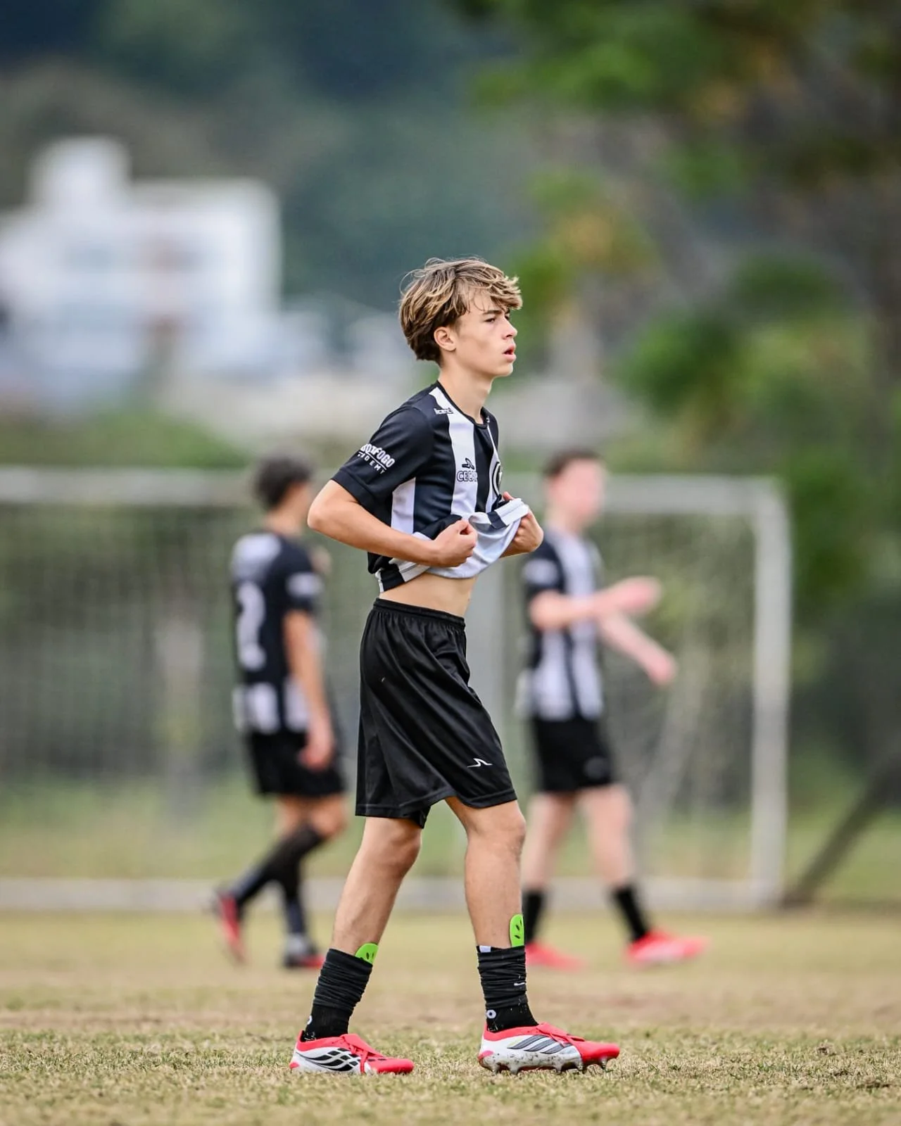 A young male soccer player in black and white uniform adjusting his jersey on a field with other players in the background.