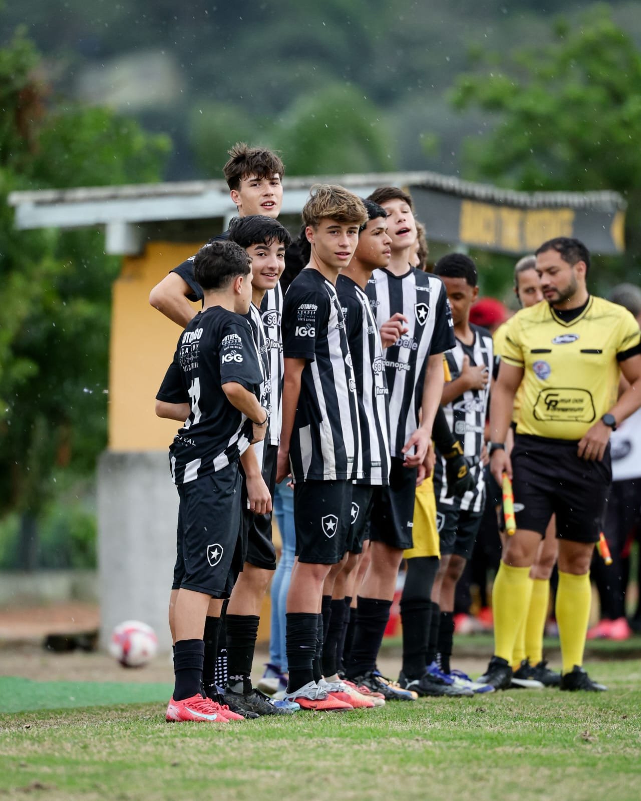 A group of young male soccer players standing in a line on a field, wearing black and white striped jerseys with the star emblem of Botafogo, with a soccer ball on the ground nearby. An official in a yellow shirt stands to the right. 