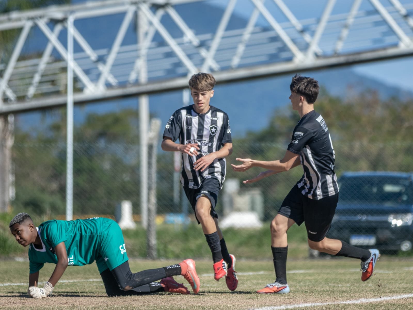 Two soccer players in black and white uniforms competing for the ball while a goalkeeper in green is on the ground, during a match on a grassy field with a fence and trees in the background.