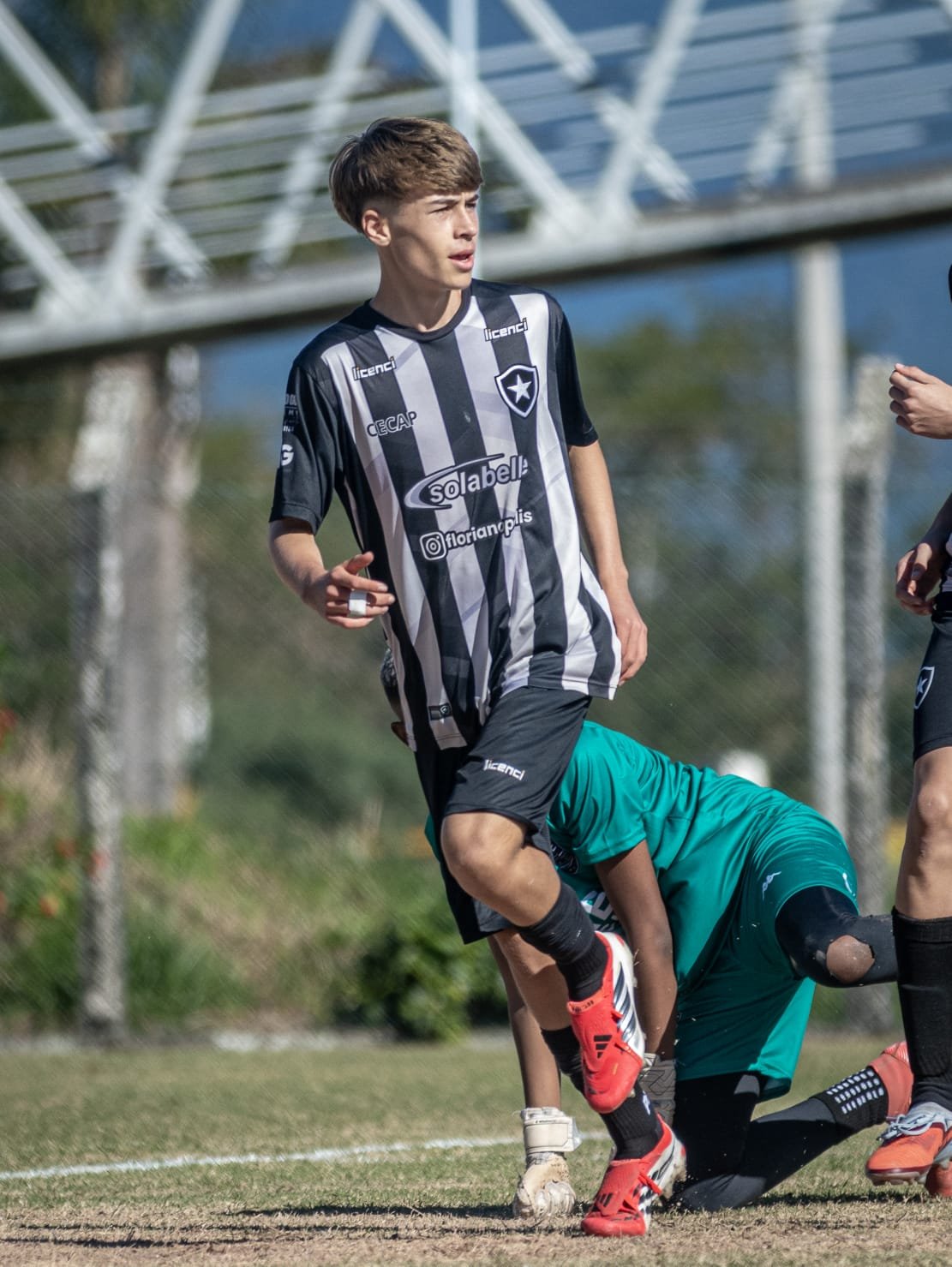 A young male soccer player in a black and white striped jersey and black shorts is on a soccer field, appearing to be in action. There is a goalkeeper in teal outfit and gloves on one knee behind him. The background includes a metal structure and tre