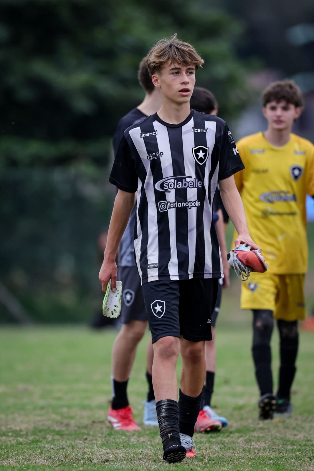 Young male soccer player with light brown hair, wearing a black and white striped jersey and black shorts, holding cleats, walking on a grassy field during a soccer game or practice.