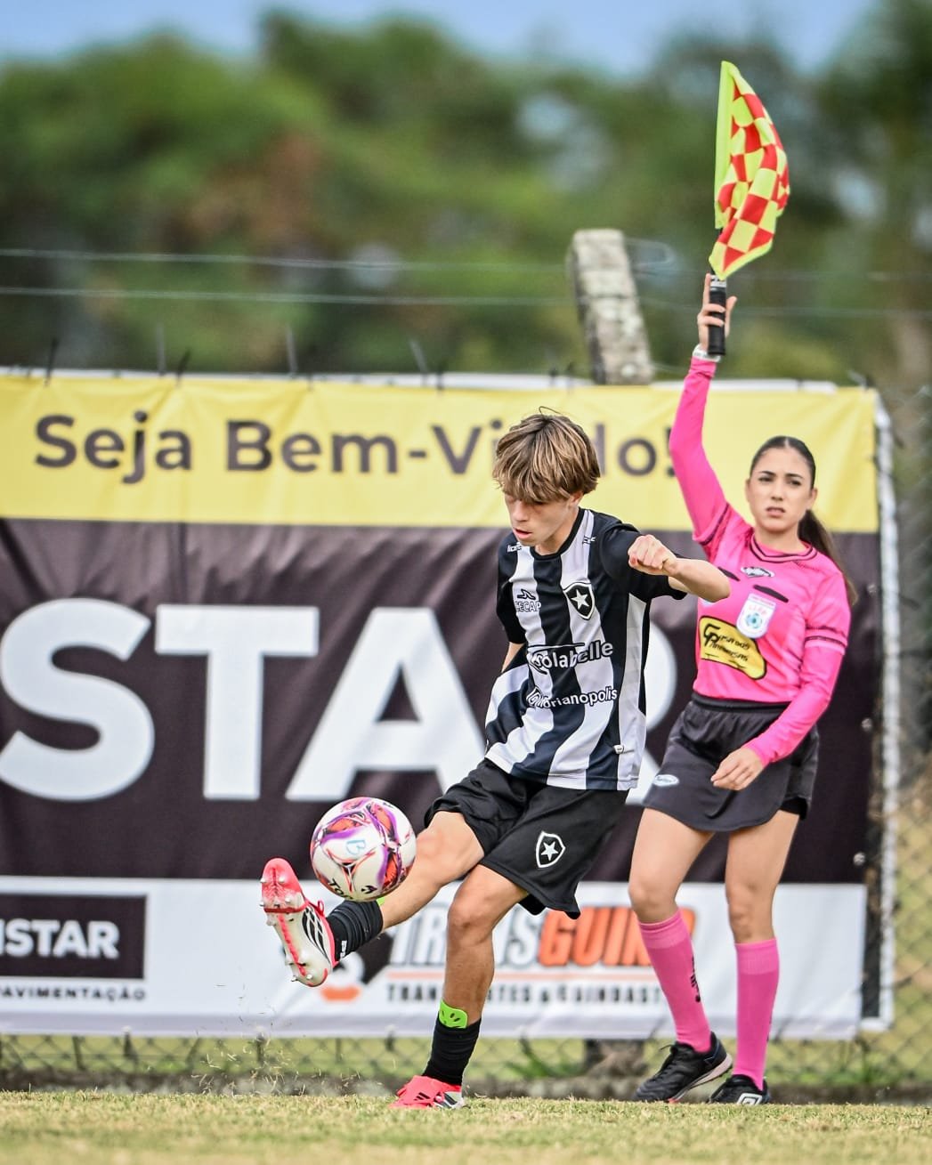 A young soccer player in a black and white uniform kicking a soccer ball while an assistant referee in a pink uniform raises her flag on a soccer field.
