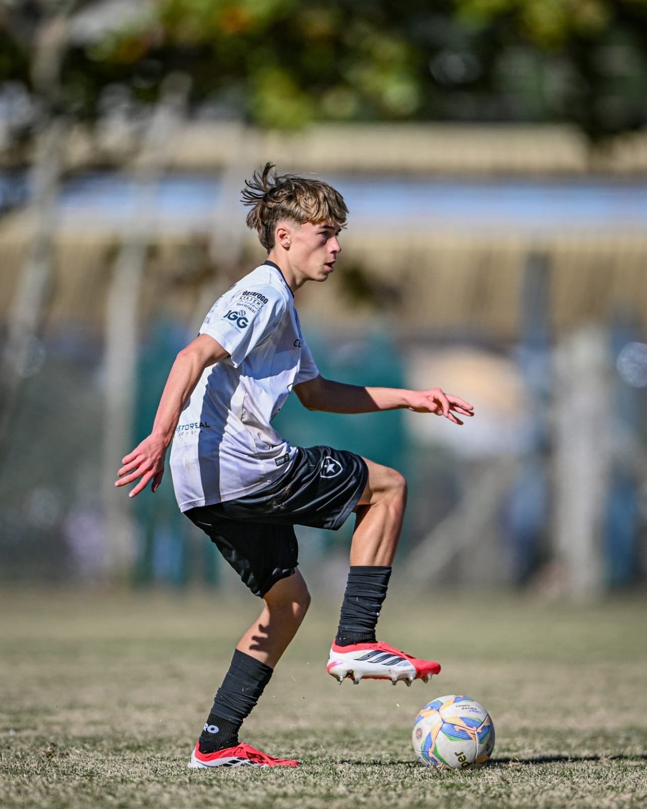 Young male soccer player in a white jersey and black shorts controlling a soccer ball on a grassy field during daytime.