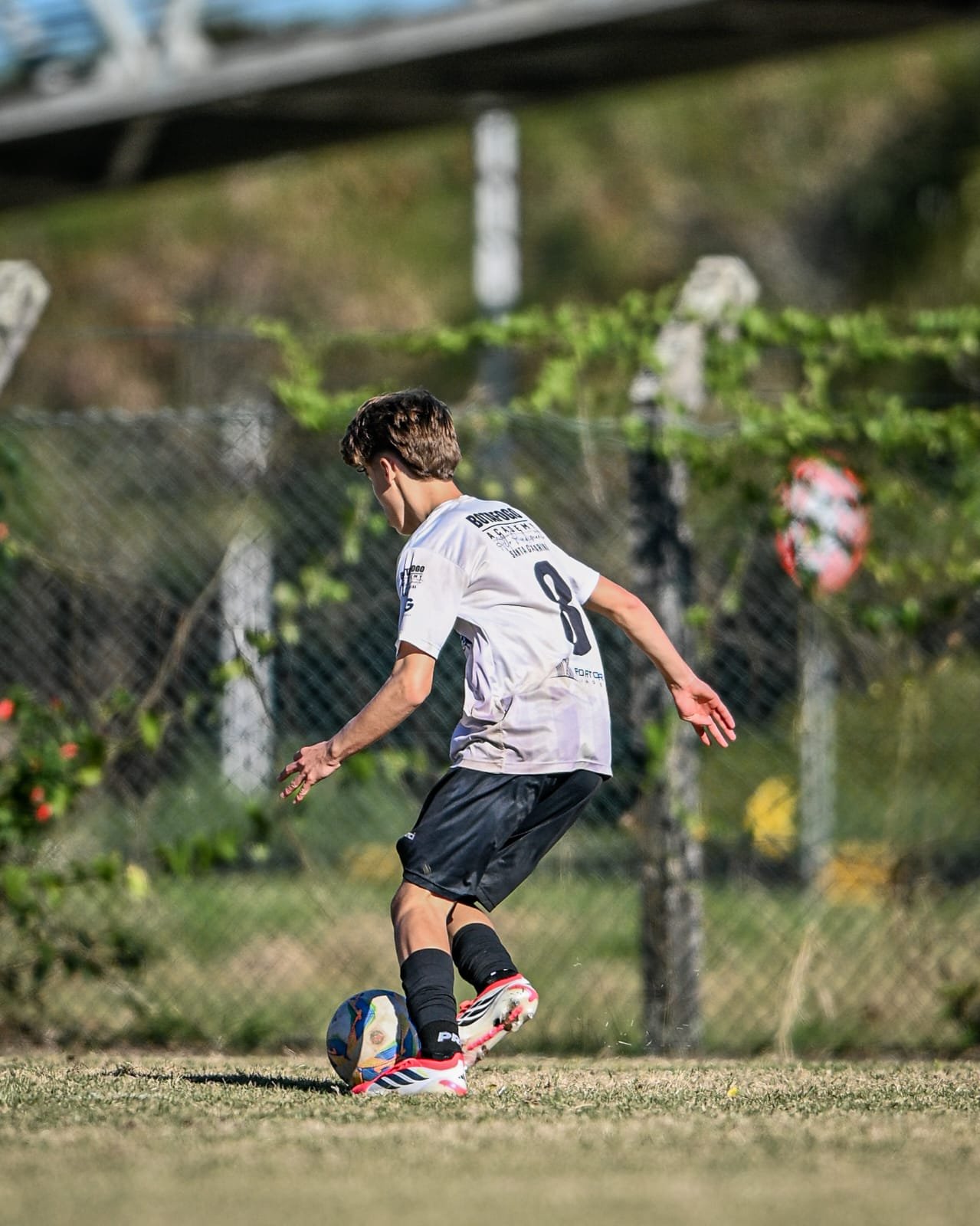 A young male soccer player in a white jersey and black shorts is preparing to kick a soccer ball on a grassy field. There is a chain-link fence and green foliage in the background.