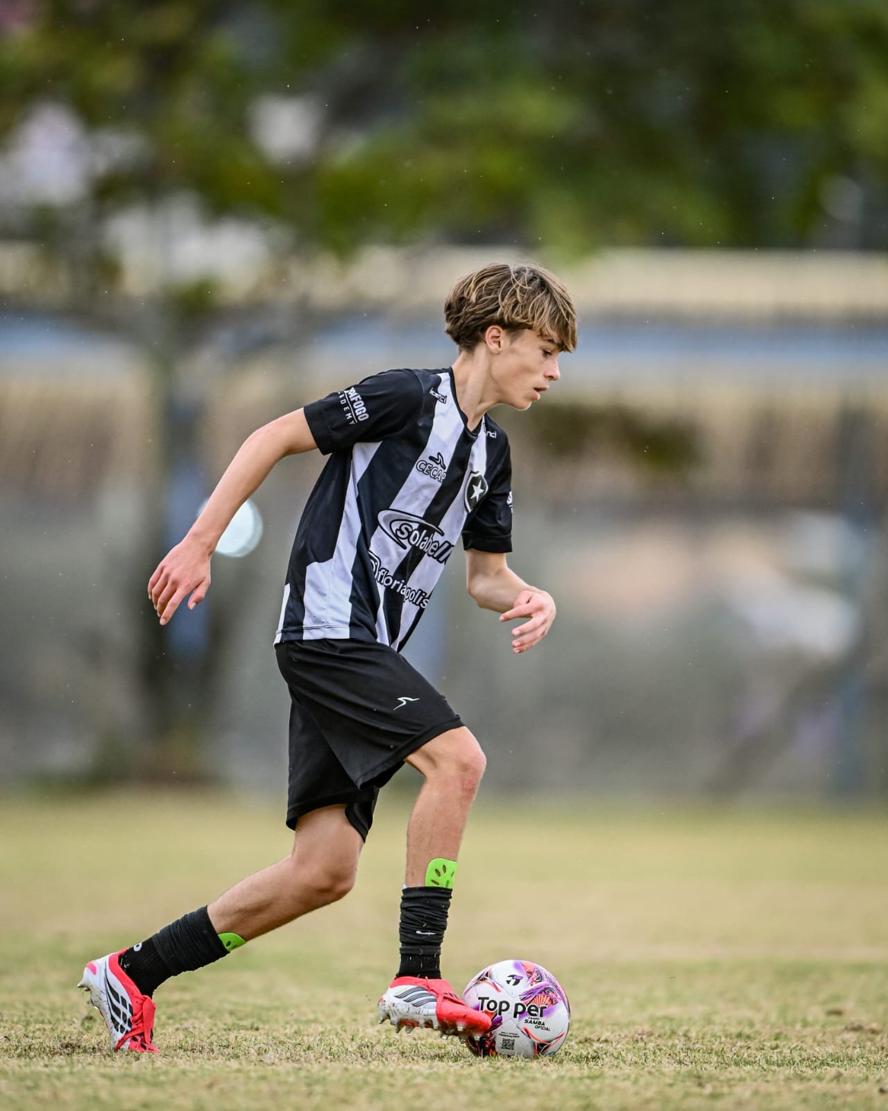 A young male soccer player wearing a black and white striped jersey, black shorts, and red soccer cleats, dribbling a pink and white soccer ball on a grassy field with trees in the background.