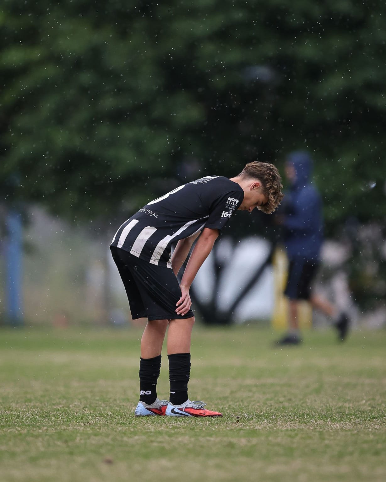 A young male soccer player for Botafogo bent over during a rainy game on a grassy field.