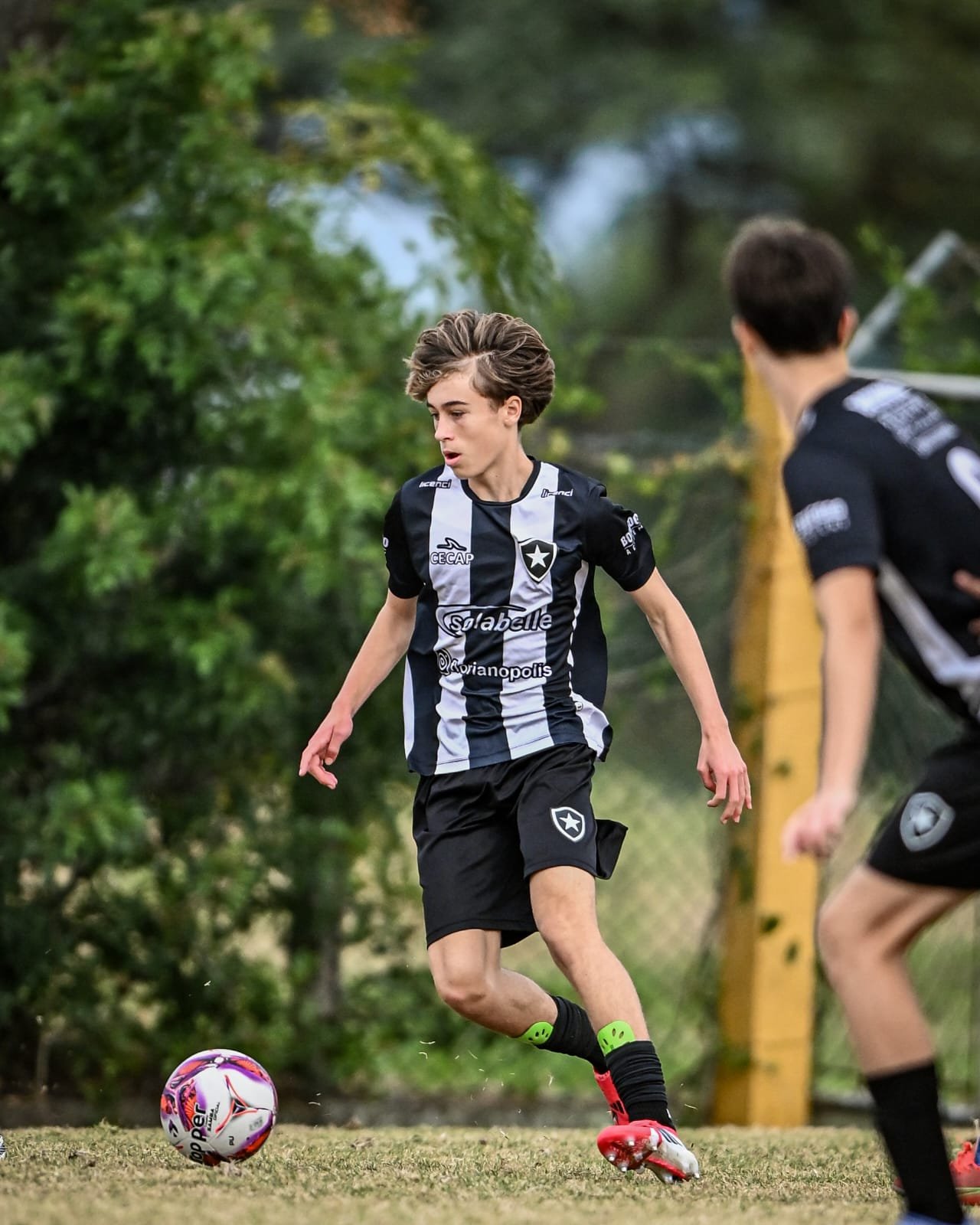 Two young soccer players in black and white striped jerseys are playing on a grassy field with trees in the background. One player is about to kick a soccer ball, wearing black shorts, black socks with green accents, and red soccer cleats.
