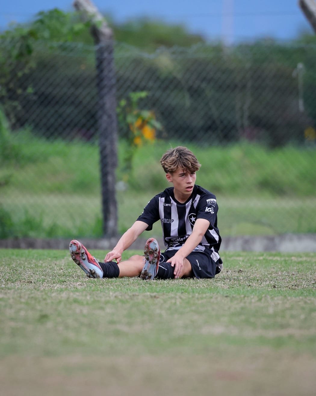 A young boy with messy hair sitting on the grass in a soccer uniform, looking distressed or tired, with his legs extended and hands resting on his knees, on a soccer field with a chain-link fence and trees in the background.