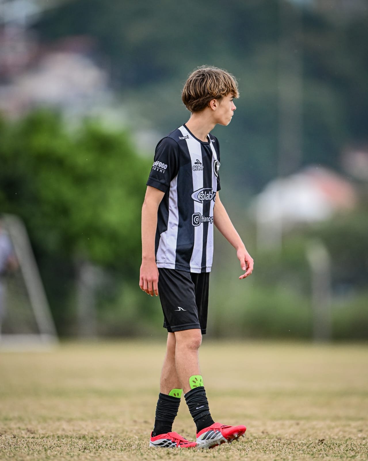 Young male soccer player standing on the field, wearing a black and white striped jersey, black shorts, black socks with green accents, and bright red cleats.