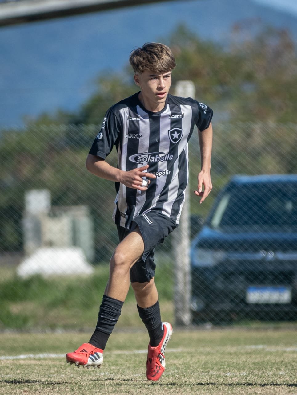Young male soccer player running on the field during a game, wearing a black and white striped uniform with logos, red cleats, and black socks.