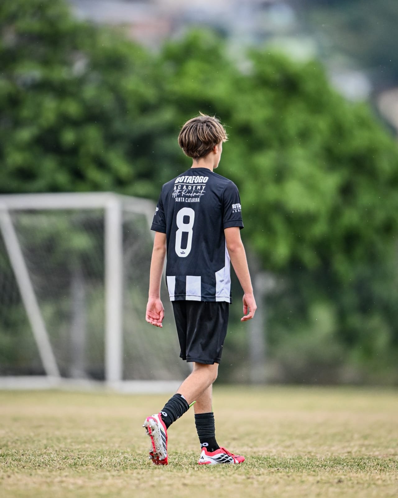 A young soccer player in a black and white jersey with the number 8, black shorts, and red and black cleats walking on a soccer field with a goalpost in the background and trees around.