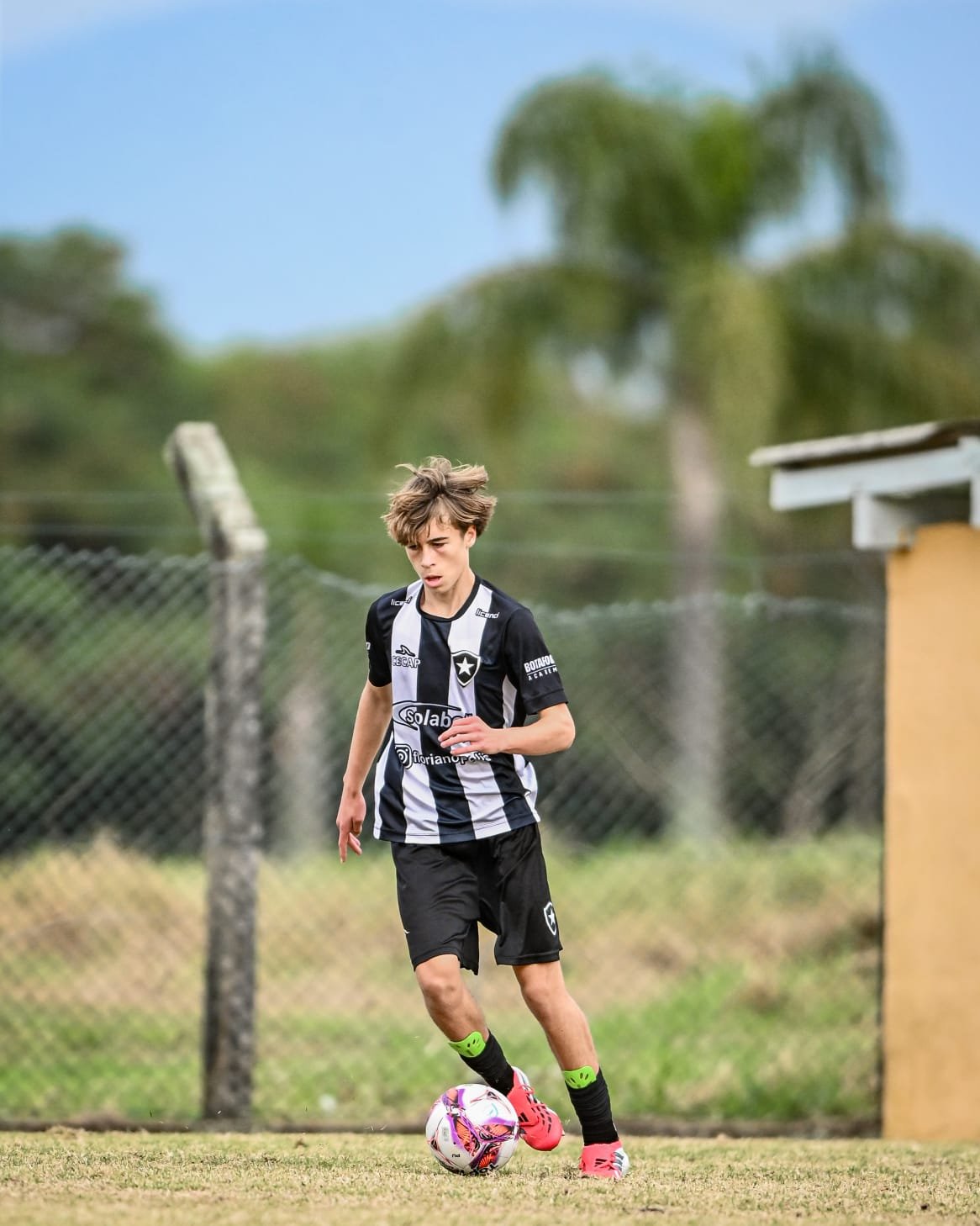 Young male soccer player in black and white uniform dribbling a soccer ball on a grassy field with a fence and trees in the background.