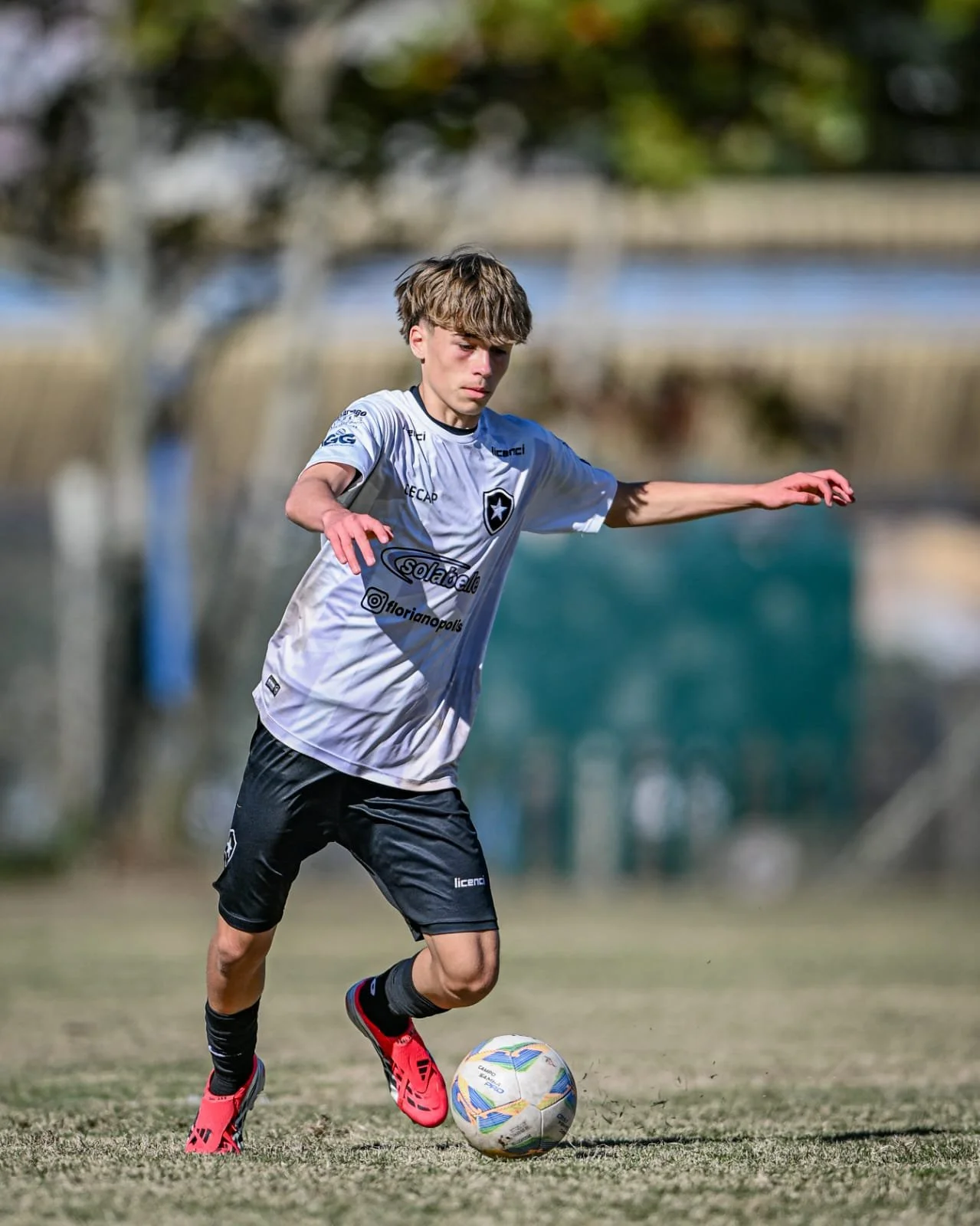 A boy playing soccer outdoors, wearing a white jersey, black shorts, red soccer cleats, and black socks, dribbling a colorful soccer ball on a grassy field with blurred trees in the background.