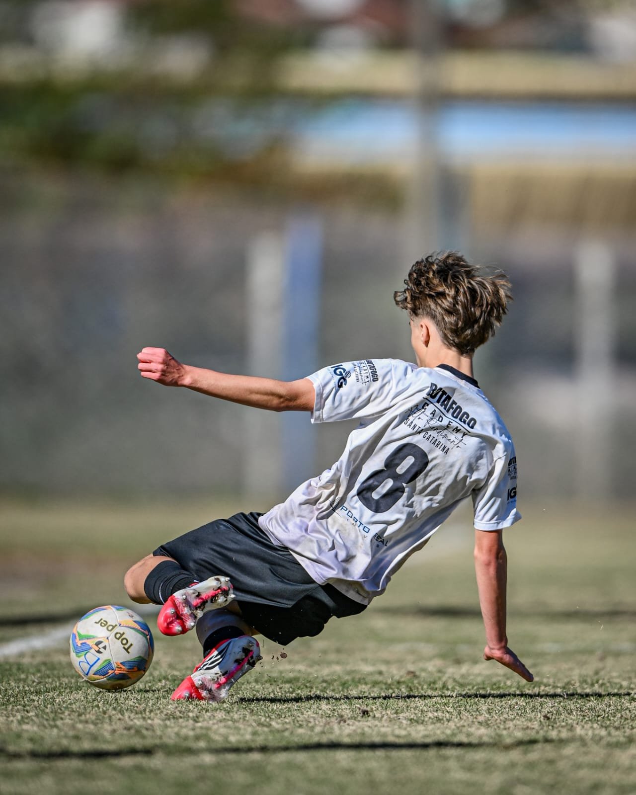 A young male soccer player in a white jersey with the number 8, sliding on the grass to kick a soccer ball during a game.