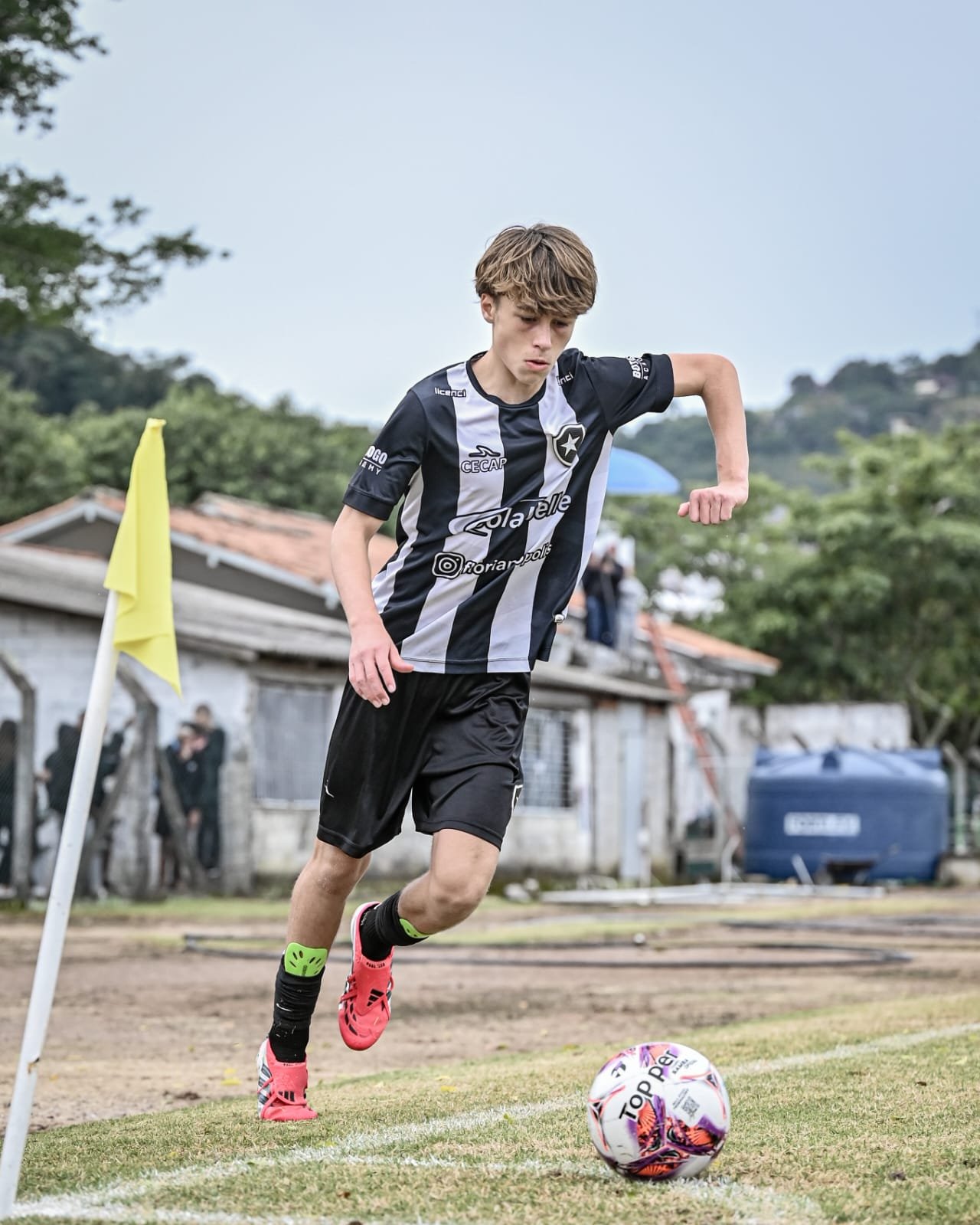 A young male soccer player in a black and white striped jersey and black shorts is kicking a colorful soccer ball on an outdoor field near a corner flag with a yellow cloth. The background shows trees, buildings, and a few spectators.