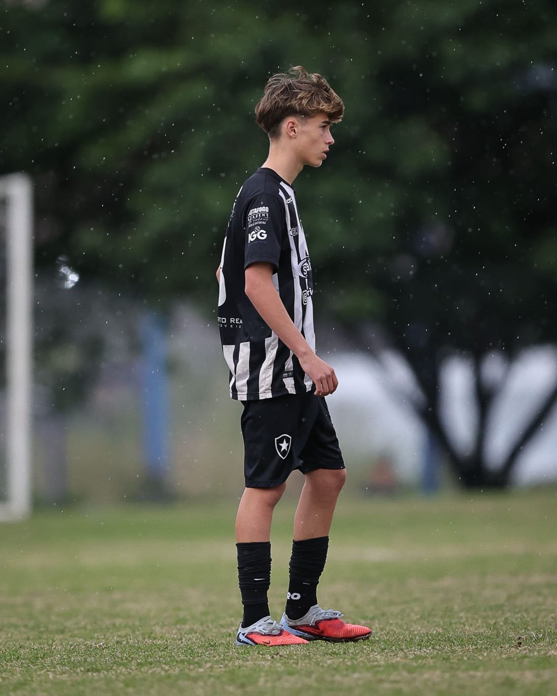 A young male soccer player stands on a grassy field during a rainy day, wearing a black and white striped soccer uniform, black socks, and red and gray cleats.