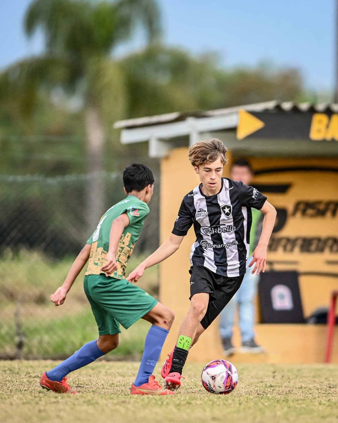 Two young boys playing soccer on a grassy field, one in a black and white striped jersey and the other in a green and gold jersey. The boy in the striped jersey is controlling the ball while the other is approaching. There is a yellow background stru