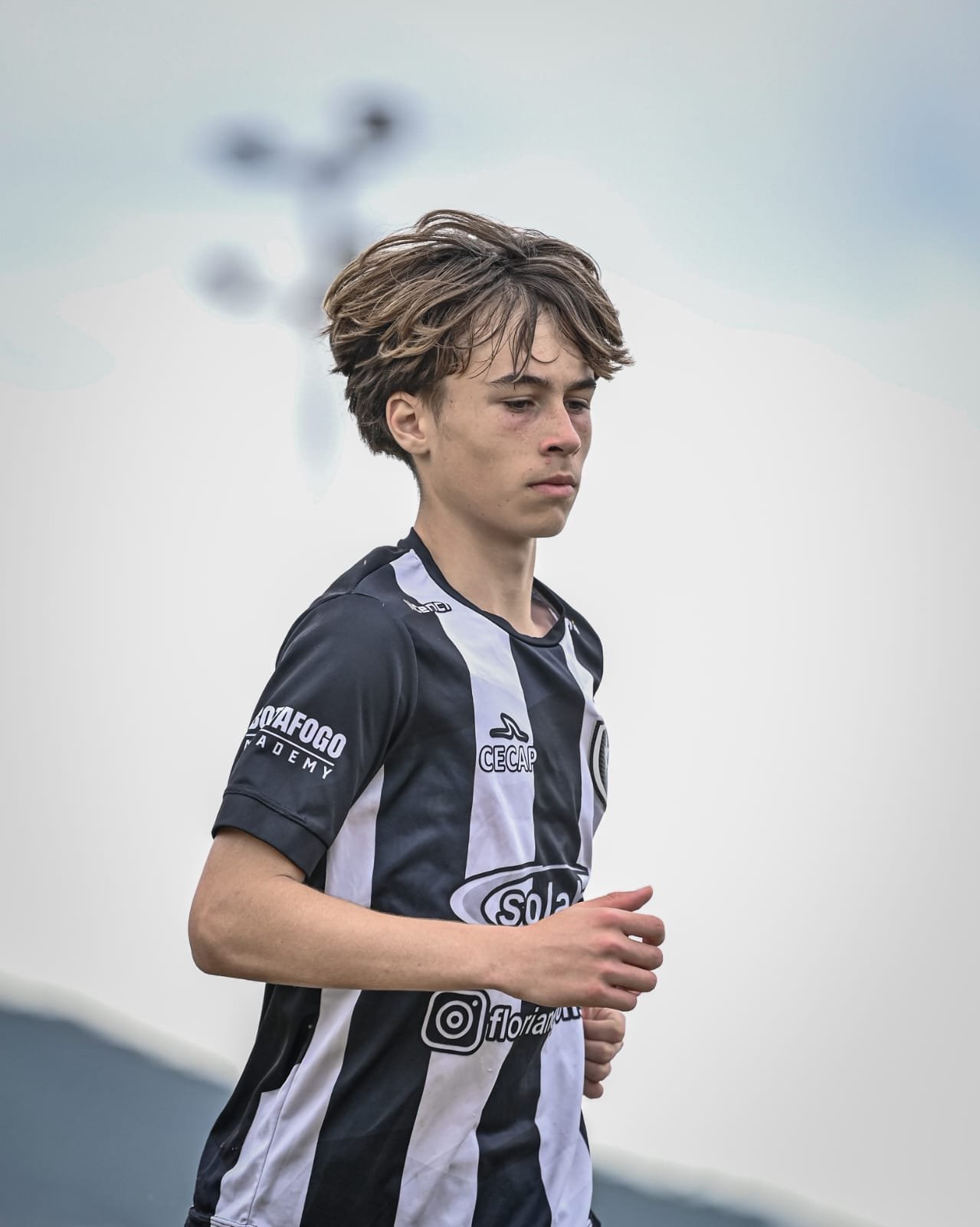 A young male athlete in a black and white striped sports jersey running outdoors with overcast sky in the background.