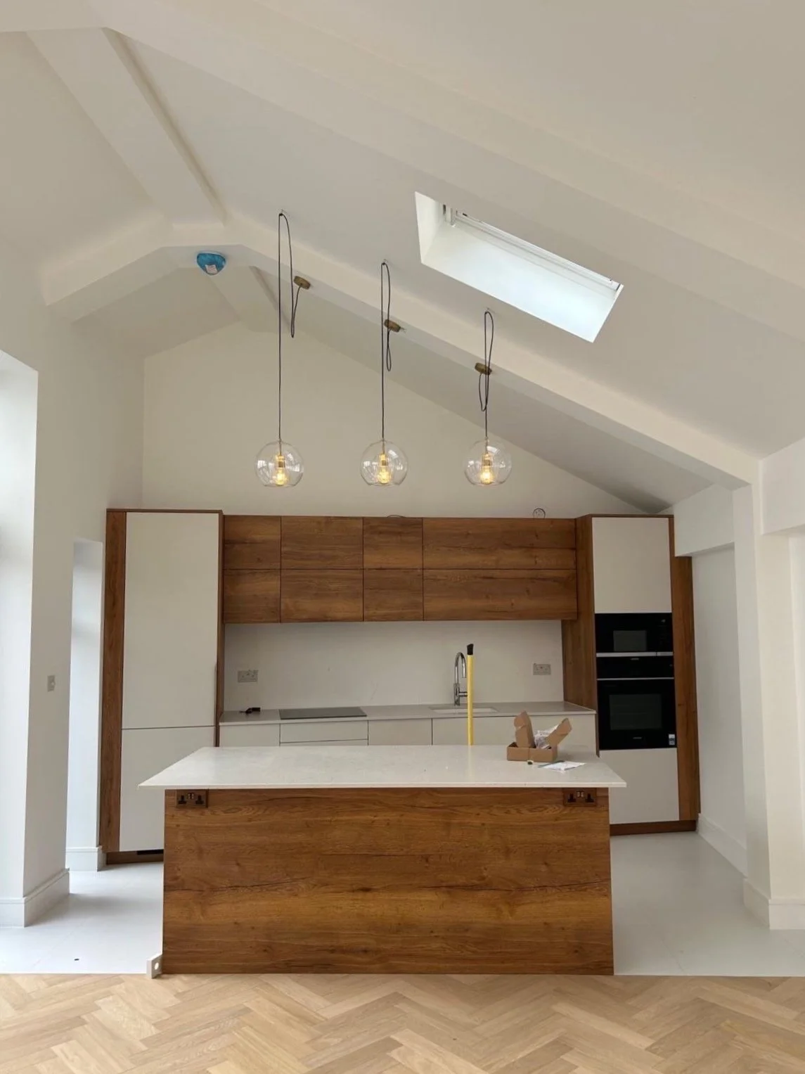 Modern kitchen with white countertops, wooden cabinets, three hanging globe pendant lights, skylight, and a built-in oven.