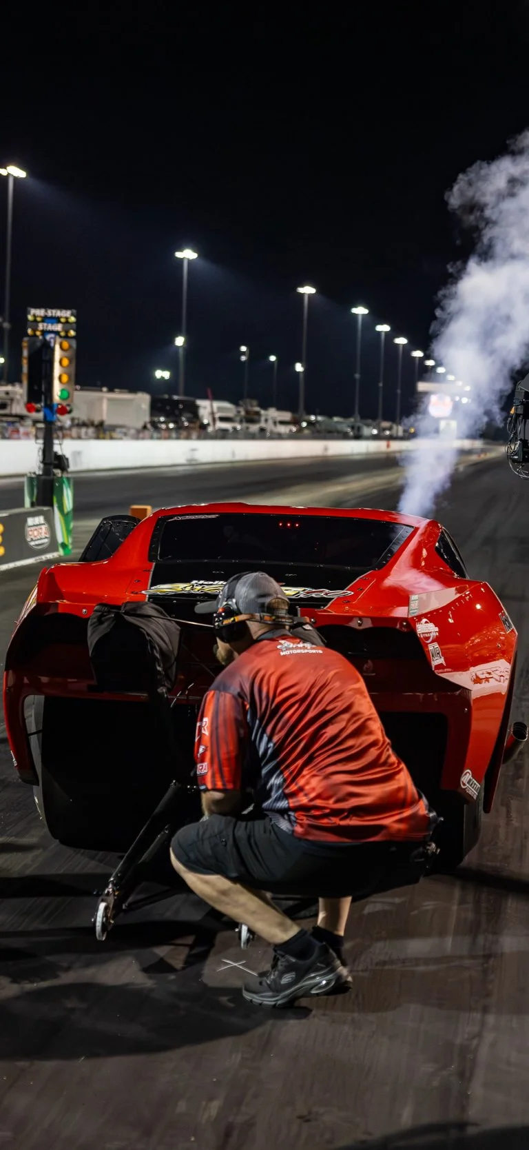 Person crouched next to a red drag racing car at night, working on it with a track in the background, smoke or steam coming from the car's exhaust.