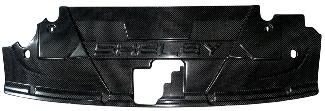 Close-up of a black carbon fiber engine cover with the word 'SHELBY' embossed in the center.