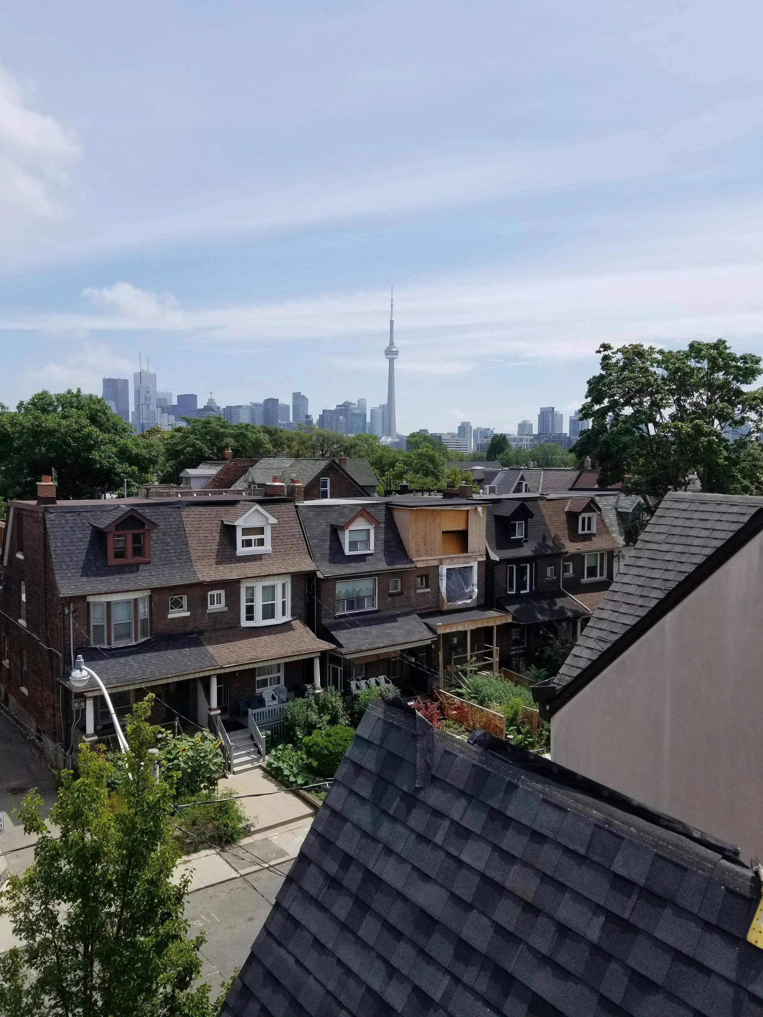View of a neighborhood with houses, trees, and a city skyline in the background, including the CN Tower in downtown Toronto, under a partly cloudy sky.