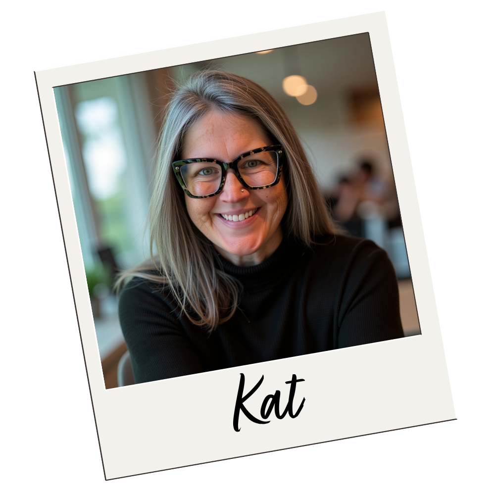 A smiling woman with shoulder-length light brown hair, wearing large tortoise shell glasses and a black top, sitting indoors near a window, with a blurred background of people and lights, and the name 'Kat' written on the photo.