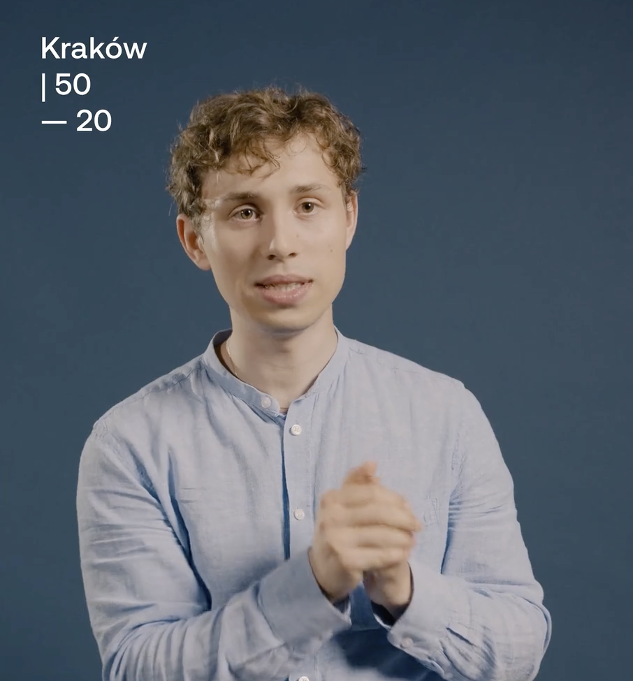 A young man with curly hair wearing a light blue shirt standing in front of a dark blue background. Text in the top left corner reads 'Kraków 50 - 20'.