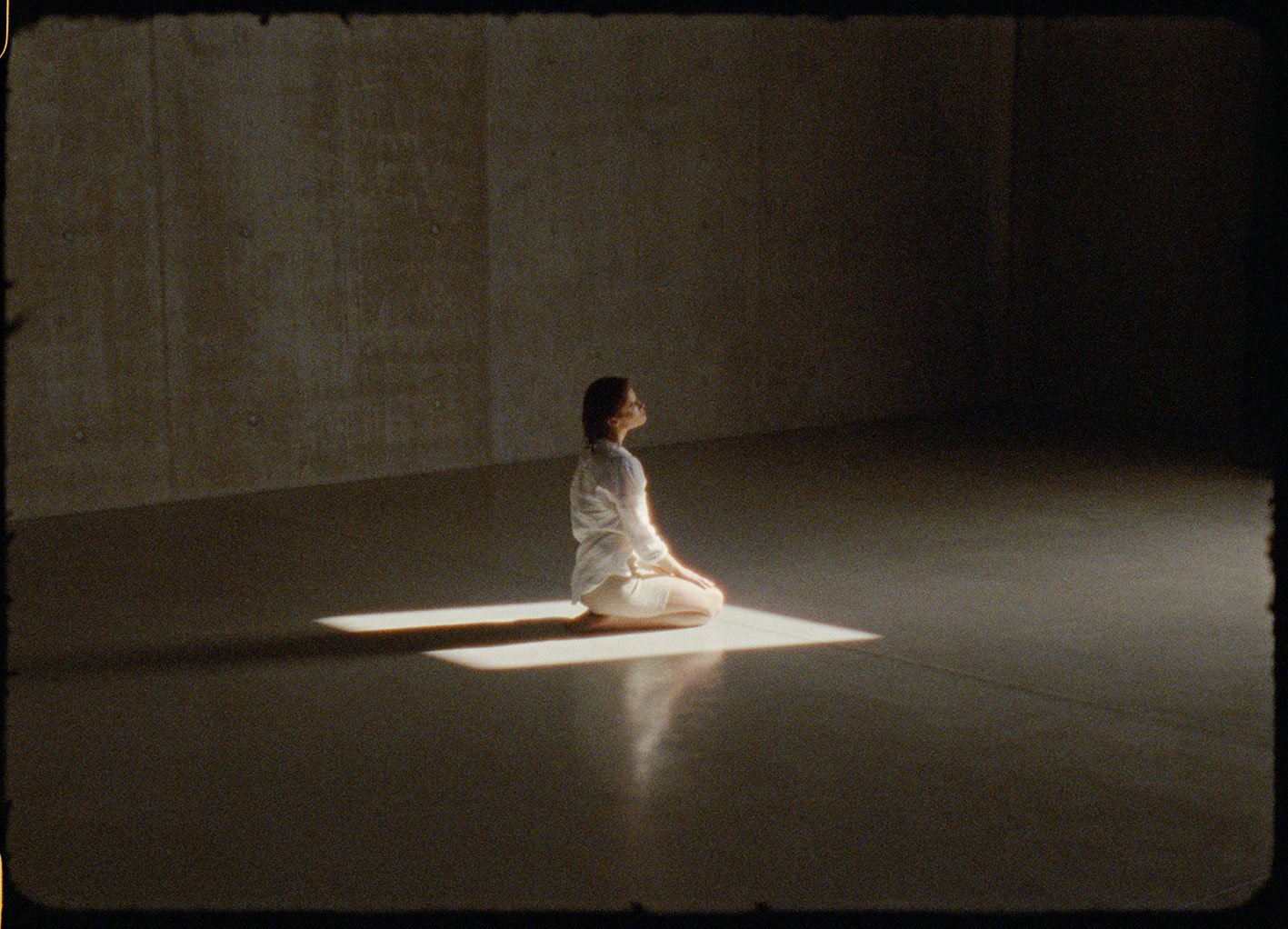 A woman practicing meditation or prayer kneeling on a small mat in a dimly lit room with a beam of light illuminating her from above.