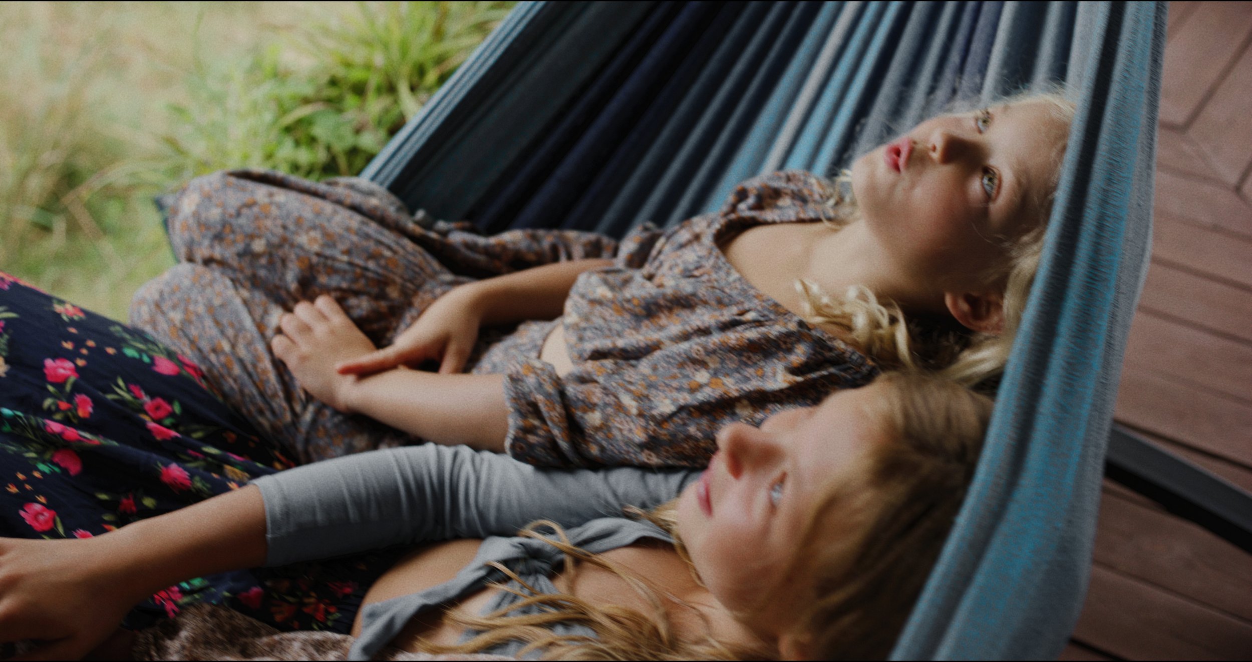 Two young girls with blonde hair lying on a hammock, looking up and relaxing outdoors.