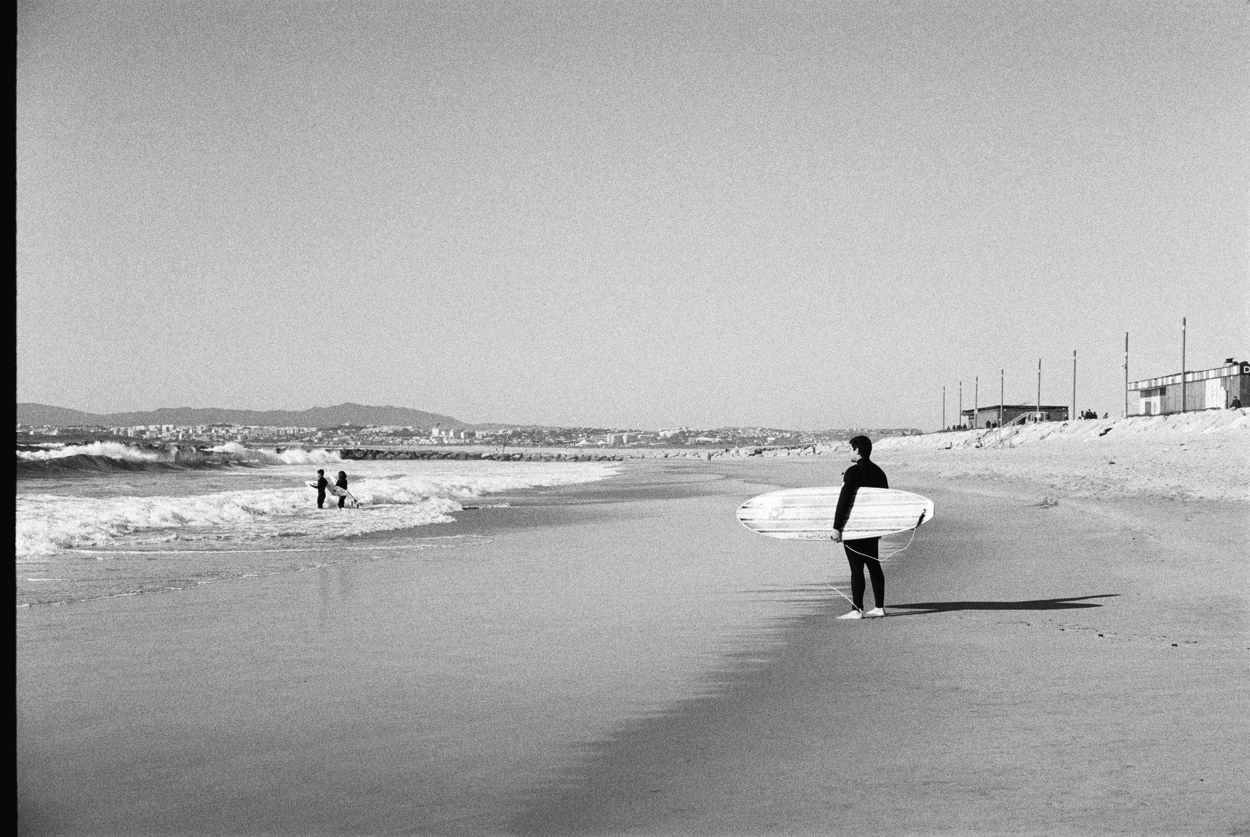 A person holding a surfboard standing on a sandy beach, looking towards two people near the water, with buildings and hills in the background.